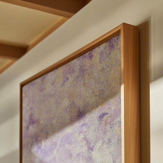 A framed abstract canvas print with a textured purple and lavender pattern, housed in a light brown wooden frame, shown hanging on a white wall.