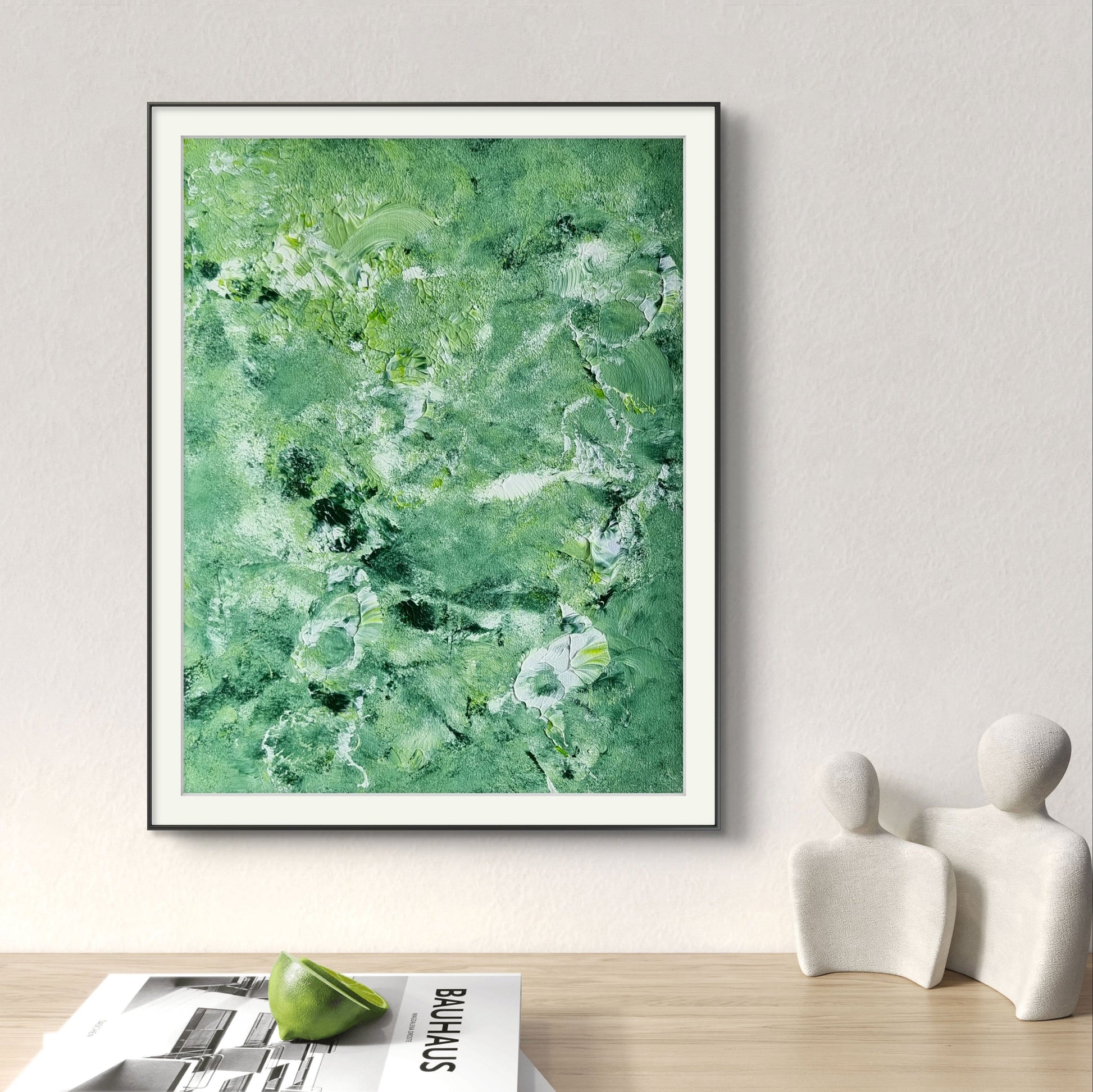 Green and white vibrant colors original, one-of-a-kind abstract painting in a black frame above an oak desk with magazines in front painted by abstract artist jeanette haga