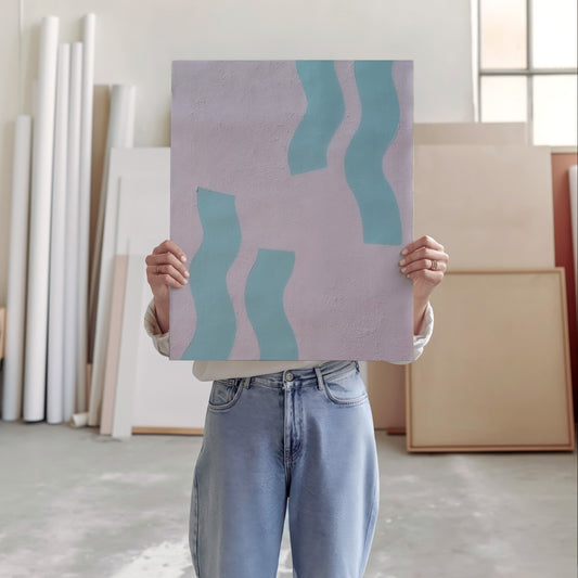 A person in a studio setting holds up a square art print or canvas with an abstract design. The print has a light lavender background with three wavy teal shapes.