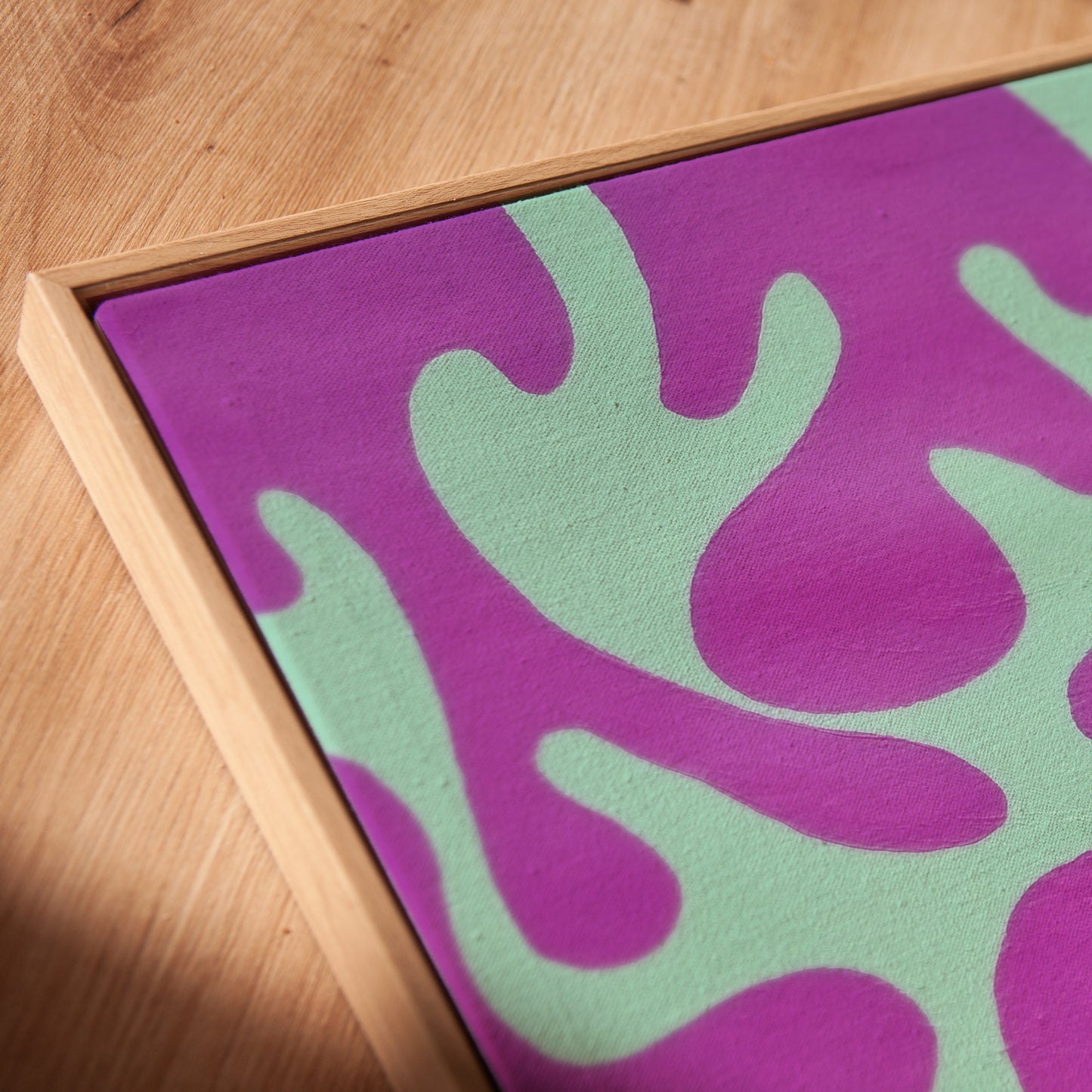 A close-up of a framed abstract art print featuring a bold purple and mint green pattern, resting on a light wood surface.