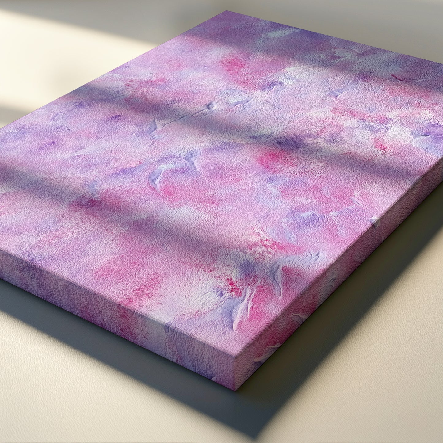 A square canvas print featuring an abstract watercolor-like pattern in shades of pink, purple, and white, displayed at an angle on a light beige surface with soft window-light shadows.