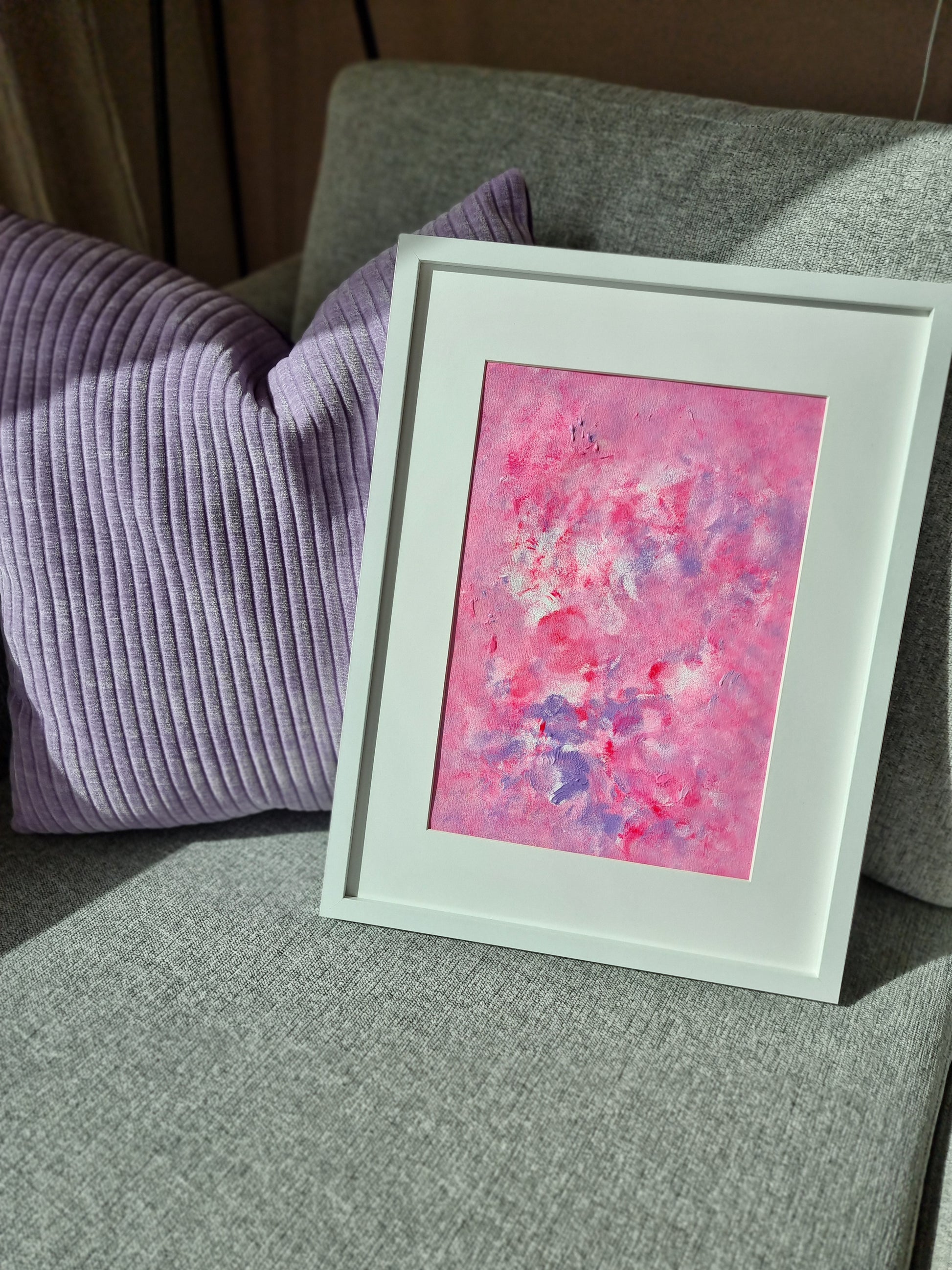 Pink, purple and white abstract original acrylic painting with a white frame and a white passe-partout leaning on a grey sofa next to a lilac cushion