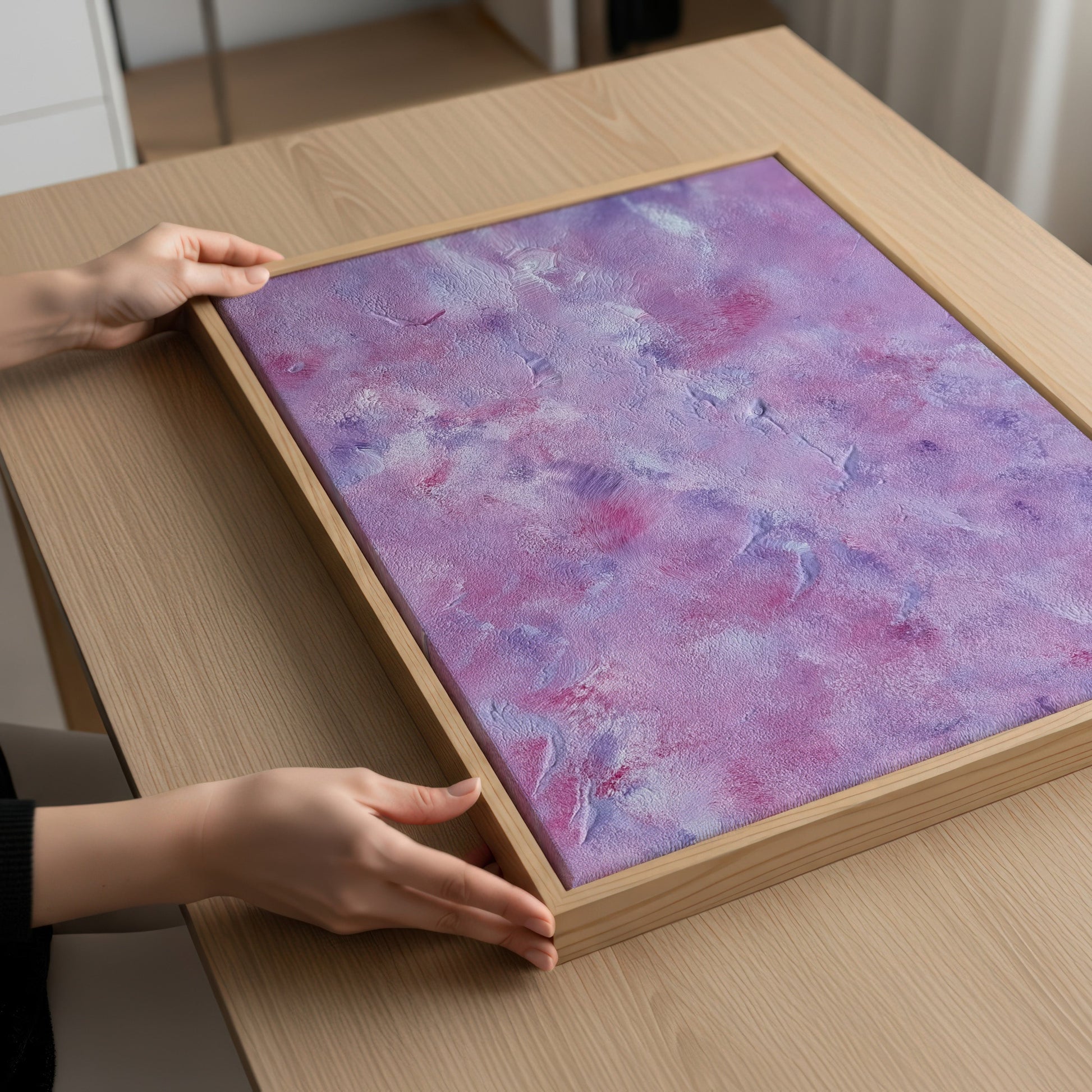 A framed abstract art print with a purple and pink marbled pattern, resting on a light wood table. Hands are visible holding the frame in place.