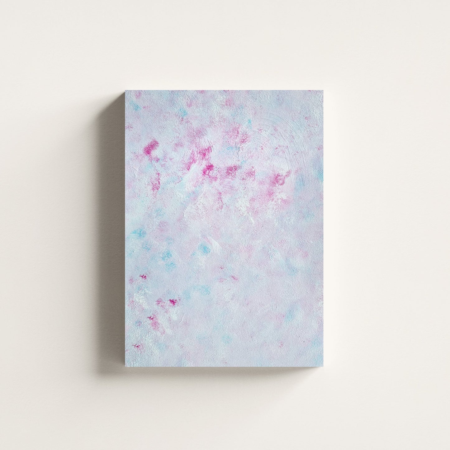 A vertical canvas wall art print featuring an abstract textured pattern of light blue, white, and pink splatters, displayed against a clean off-white wall.