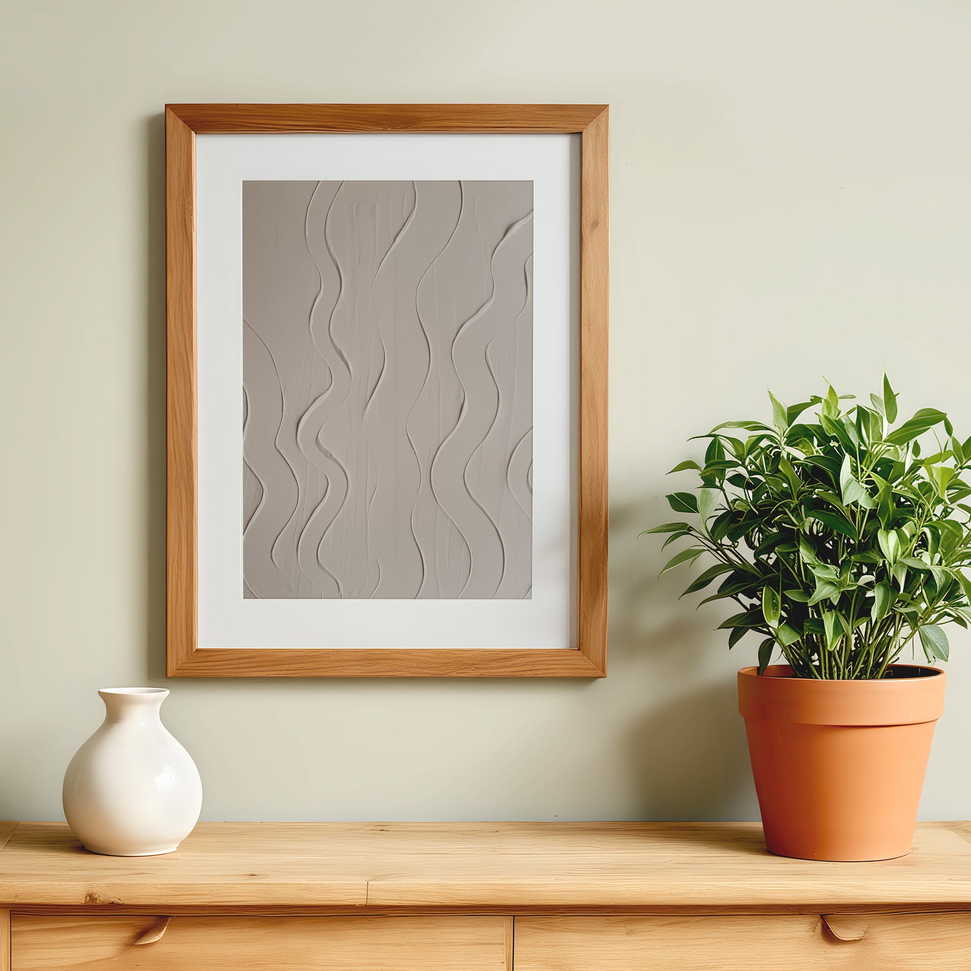 A framed abstract art print with a textured gray and beige wavy pattern, displayed on a light beige wall above a wooden console table with a white ceramic vase and a potted green plant.