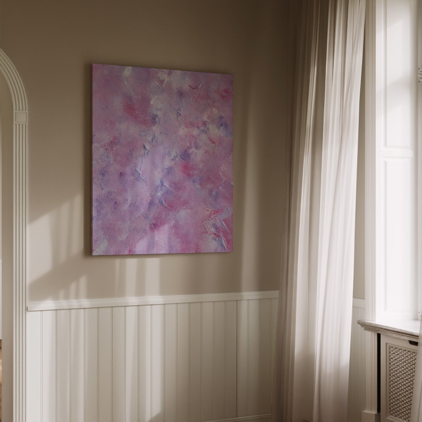 An abstract canvas painting with pink, purple, and blue tones hanging on a beige wall in a sunlit room with white wainscoting and sheer curtains.