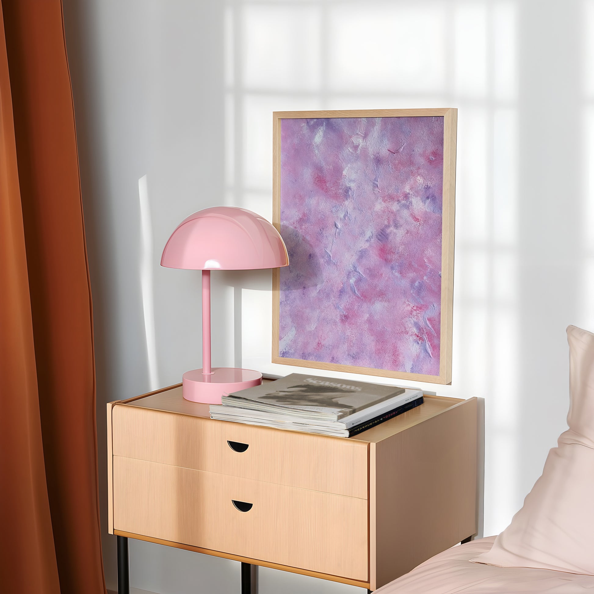 A framed abstract art print with purple and pink tones hangs on a light gray wall above a light wood nightstand. A pink dome lamp and stacked magazines sit on the table, with a rust-colored curtain on the left and a bed corner on the right.