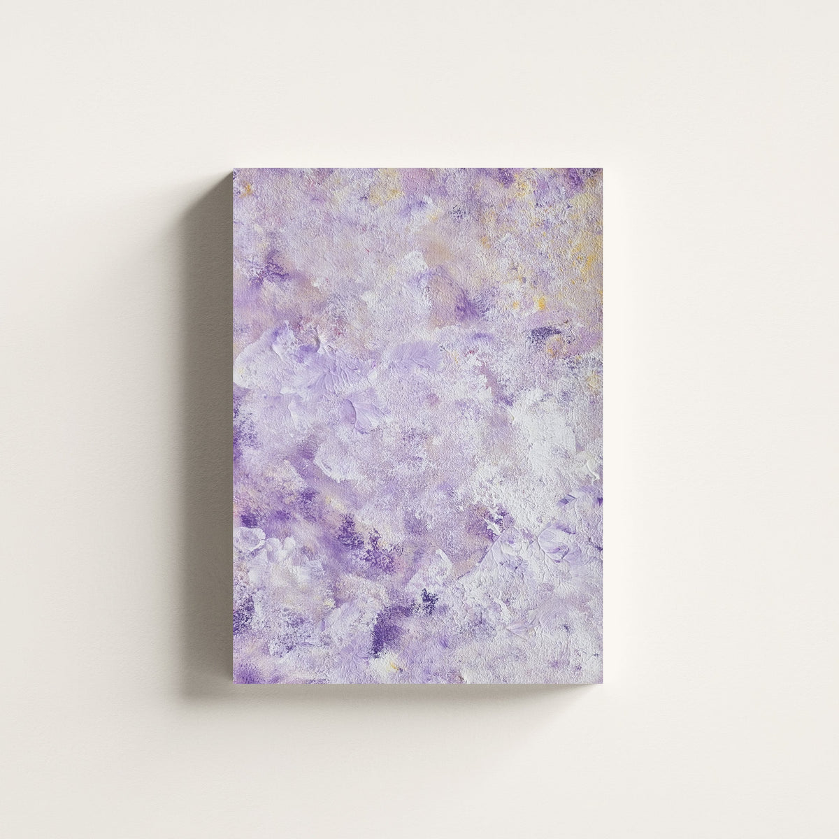 A vertical canvas wall art print featuring an abstract textured pattern in shades of lavender purple, white, and soft gray, displayed against a clean off-white wall.