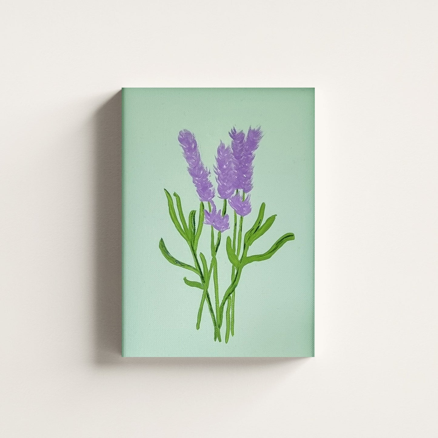 A vertical canvas wall art print featuring a painted lavender bouquet in purple and green on a pale mint-green background, shown hanging against a light neutral wall.