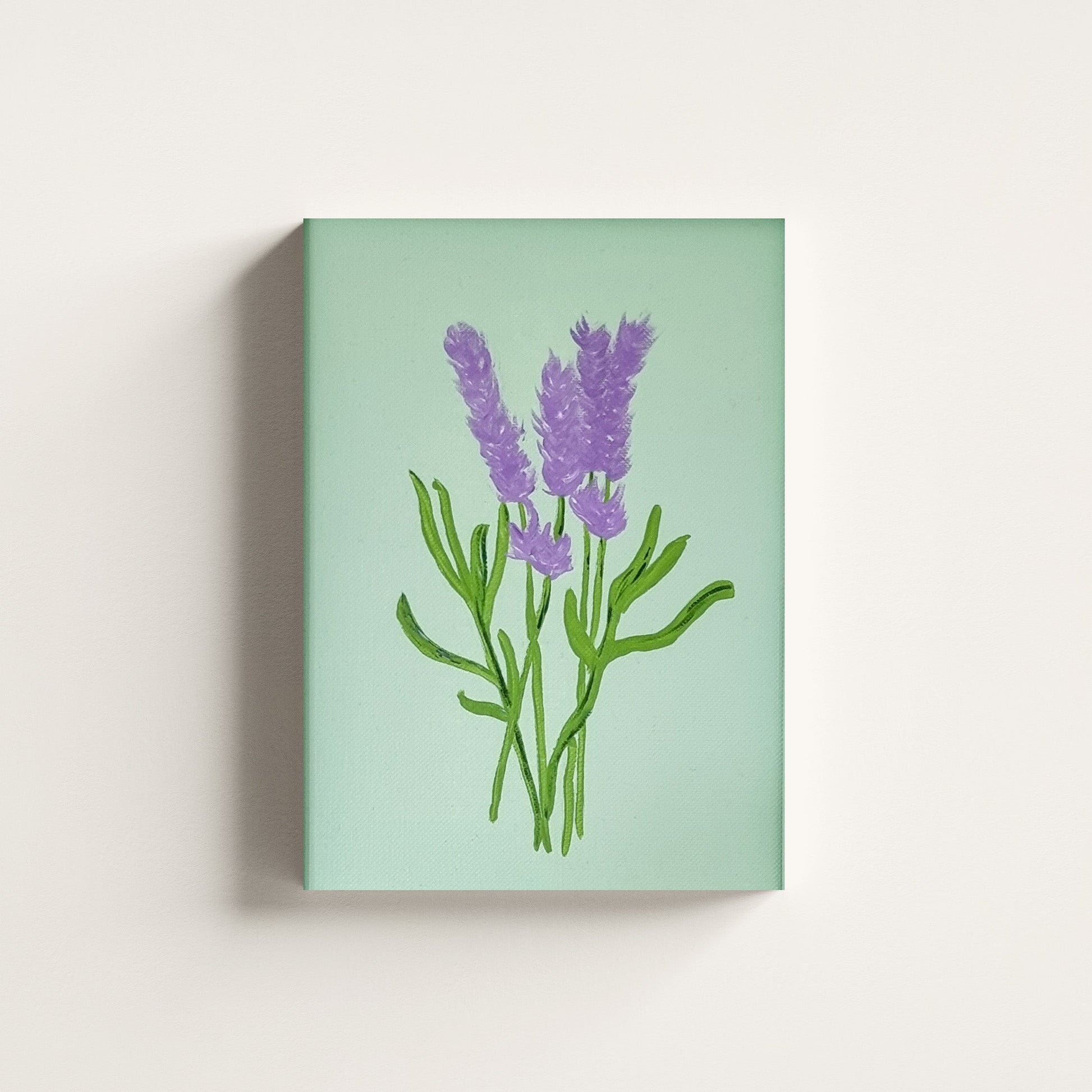 A vertical canvas wall art print featuring a painted lavender bouquet in purple and green on a pale mint-green background, shown hanging against a light neutral wall.
