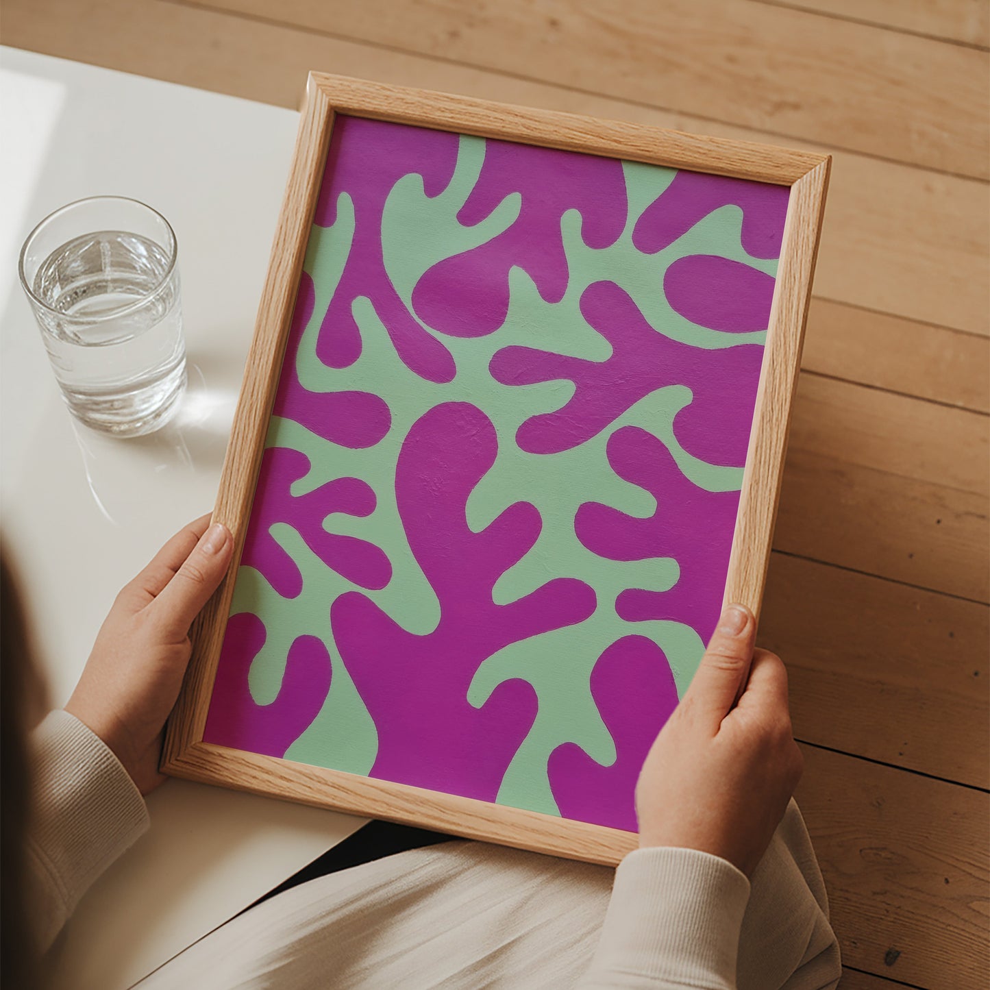 A person holds a framed abstract art print featuring a bold purple and mint green organic pattern, set against a warm wood floor background.