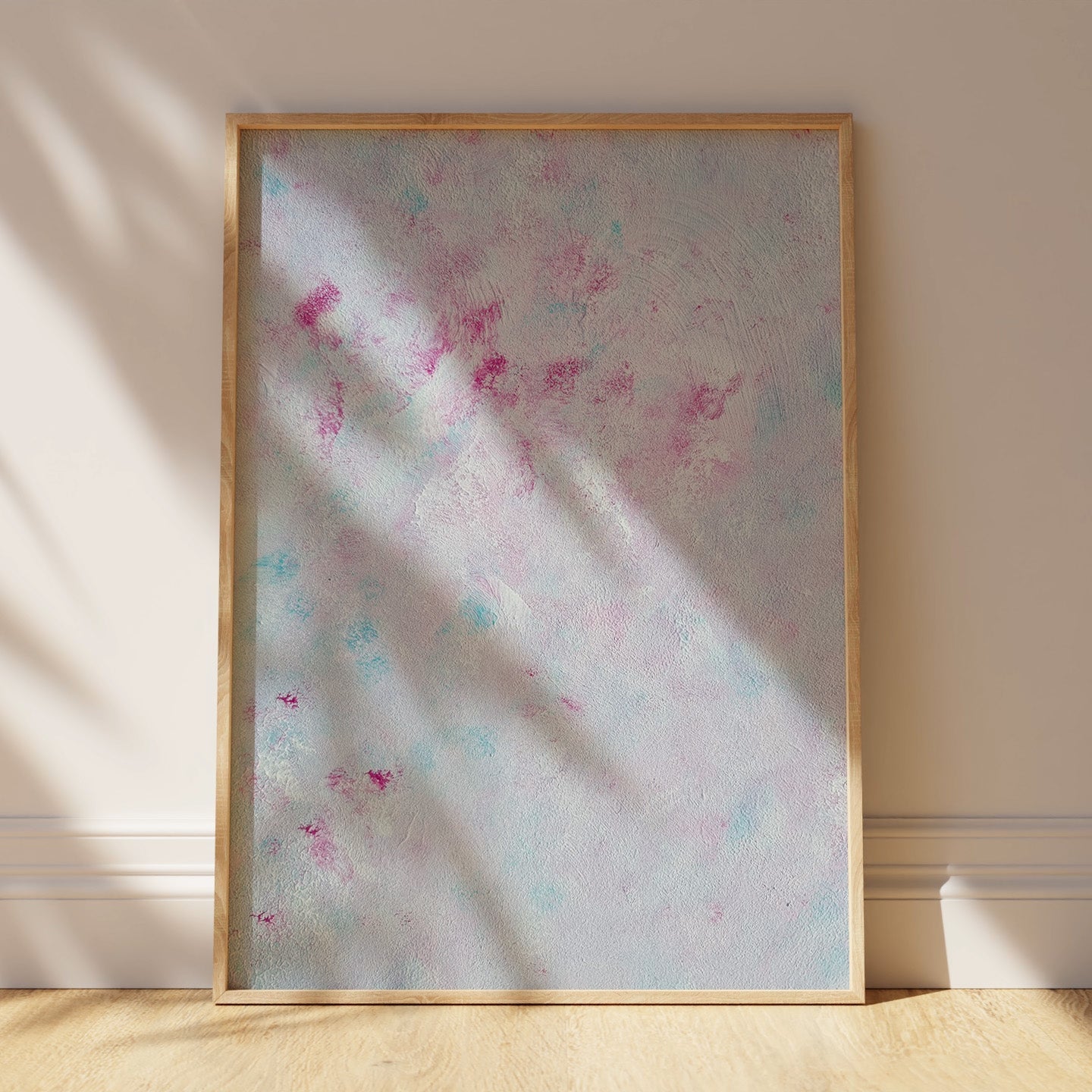 A framed abstract art print leaning against a beige wall on a light wood floor, with soft sunlight casting leaf shadows across the artwork and wall.