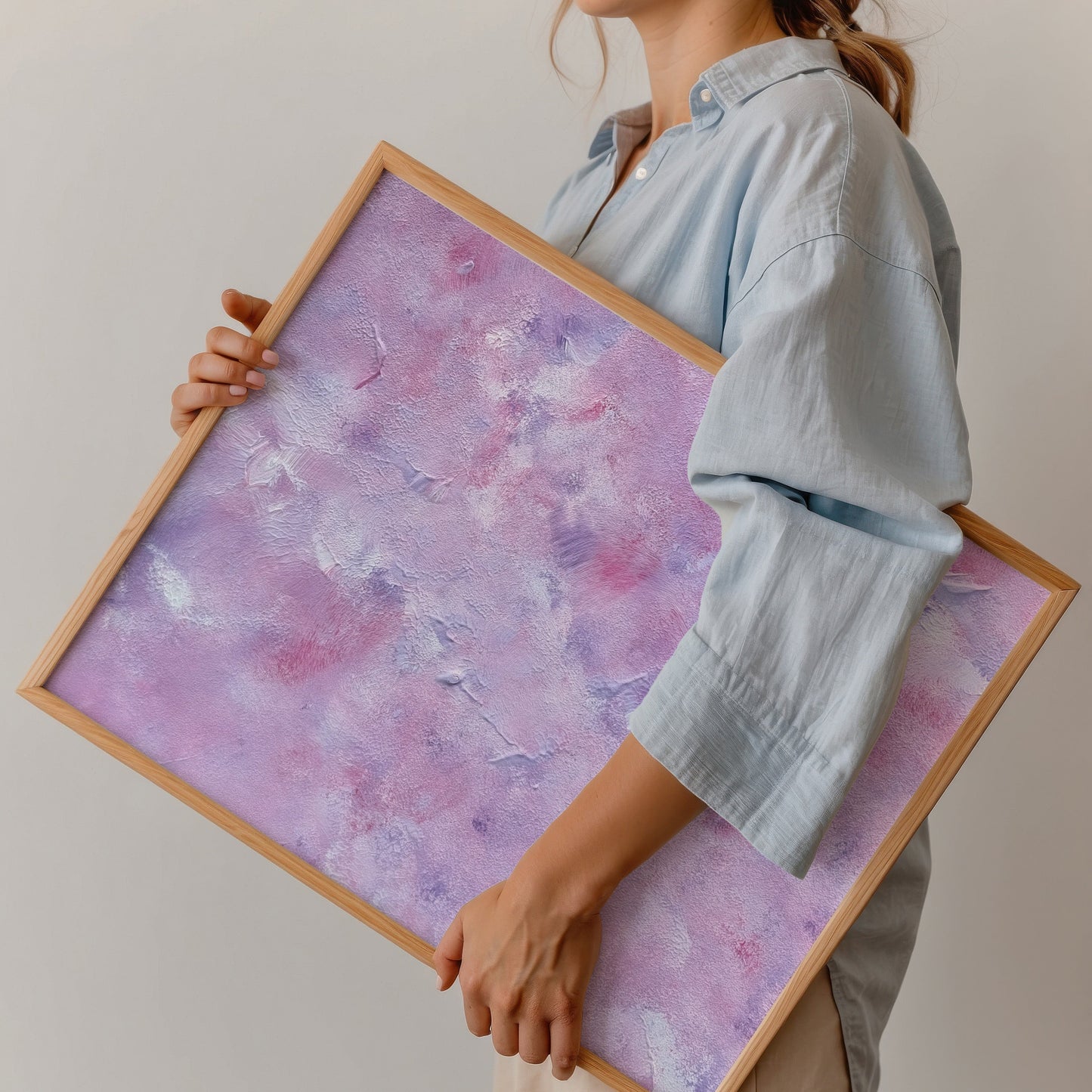 A person holding a large framed abstract art print featuring a textured purple and pink design with a light wood frame, wearing a light blue shirt and beige pants against a plain white wall.