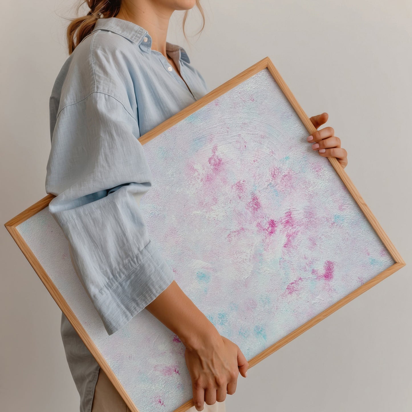 A woman in a light blue shirt and beige pants holds a large square framed art print. The print features a textured, abstract design in pastel pink, purple, and light blue on a white background, enclosed in a natural wood frame.