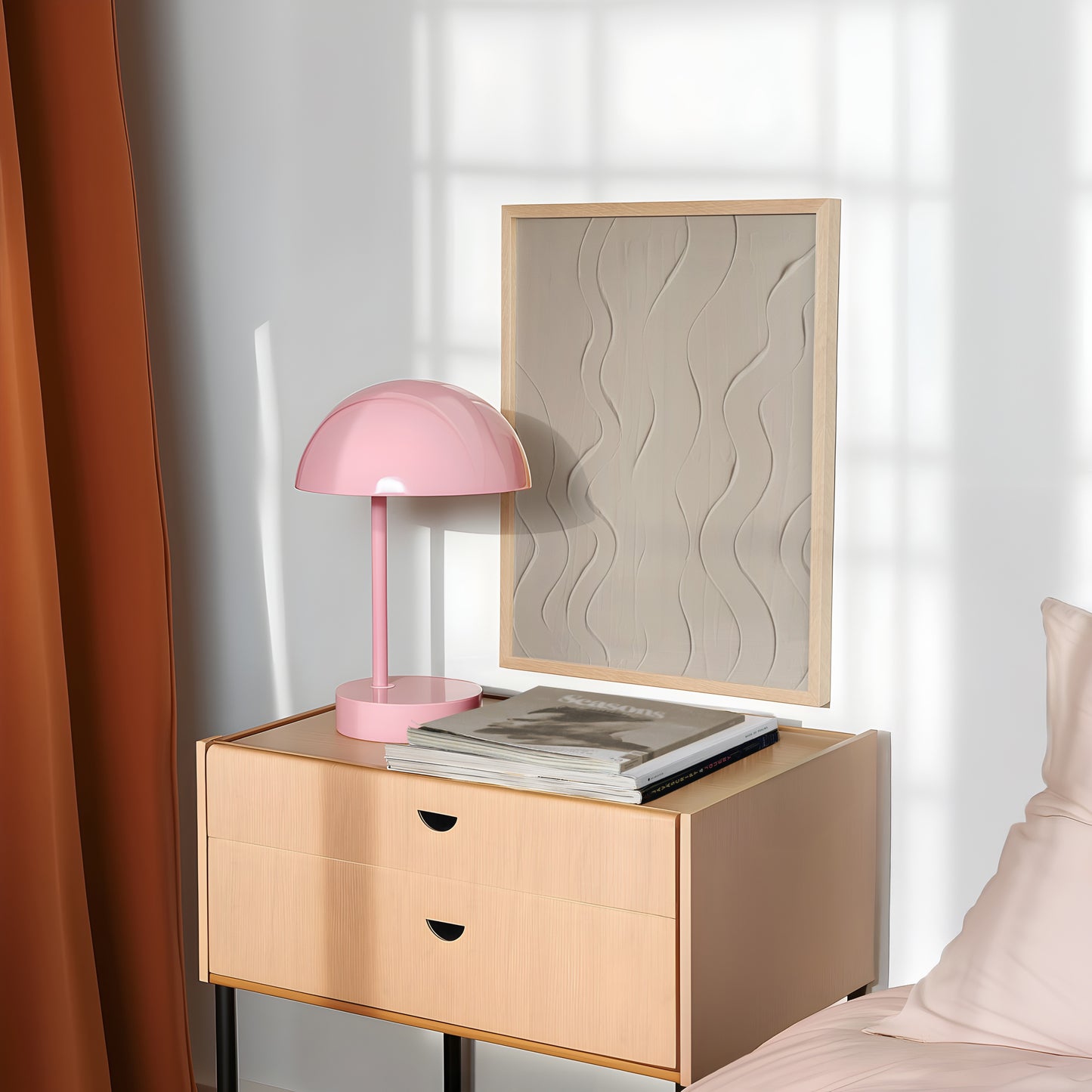 A light wood nightstand in a bedroom setting, styled with a pink dome lamp and a stack of magazines, next to a bed with pink bedding and a textured beige framed print on the wall.