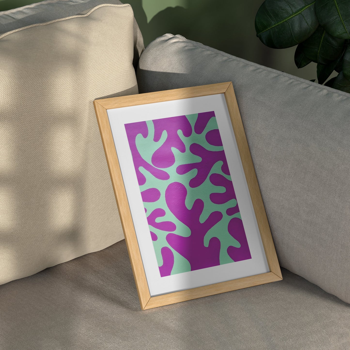 A framed abstract art print with a purple and mint green pattern, set in a light wood frame leaning against a gray sofa with a beige pillow and a green plant in the background.
