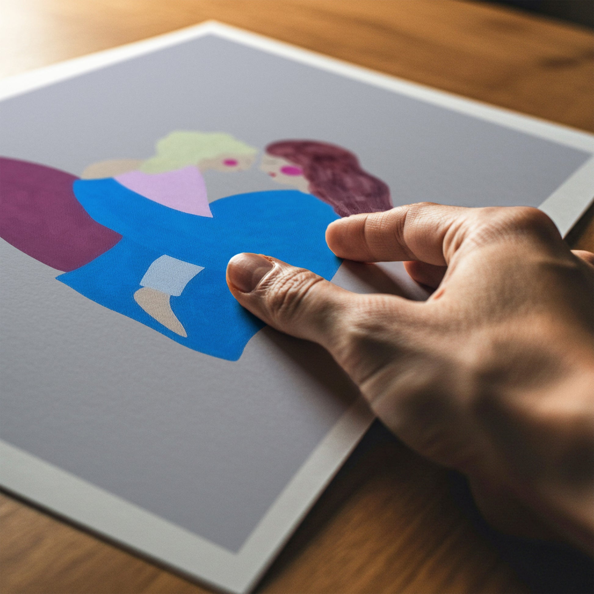A hand is holding a square art print featuring a colorful abstract illustration of two figures, set against a warm wooden background.