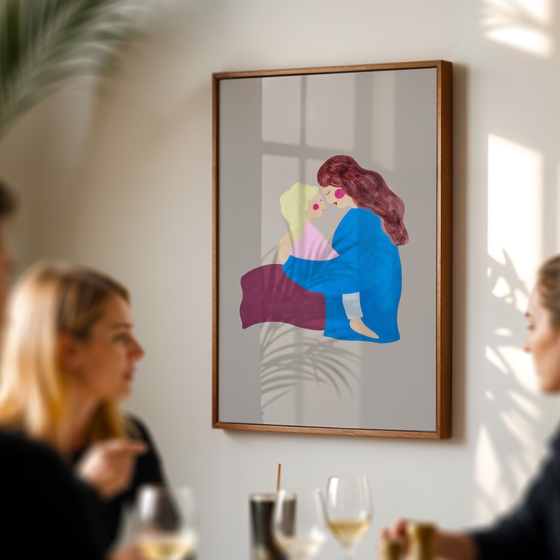 A framed art print featuring a colorful illustration of a woman in a blue dress and red hair hugging a child, displayed on a light gray wall in a dining room setting with blurred people and wine glasses in the foreground.