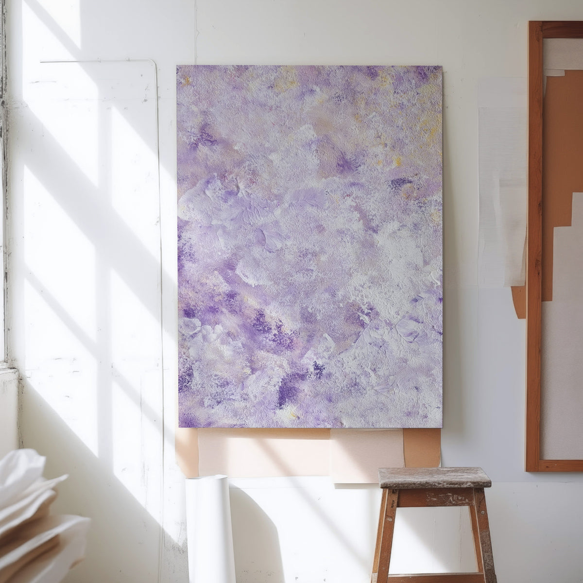An abstract canvas painting with textured purple, lavender, and white brushstrokes hanging on a white wall in a sunlit studio space, with a wooden stool below and rolled paper nearby.