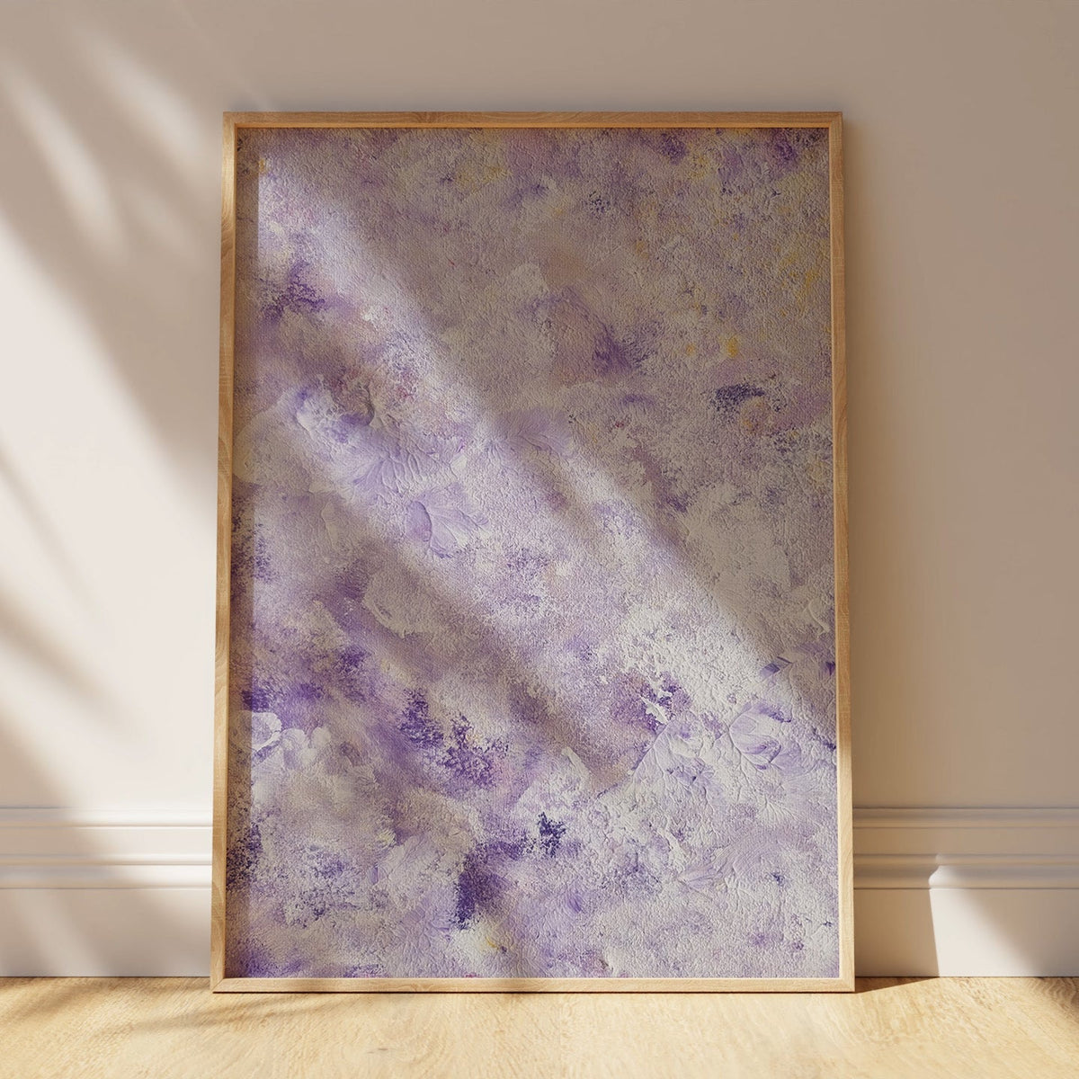 A framed abstract art print with textured purple, lavender, and cream brushstrokes, leaning against a beige wall on a light wood floor with soft window shadows.