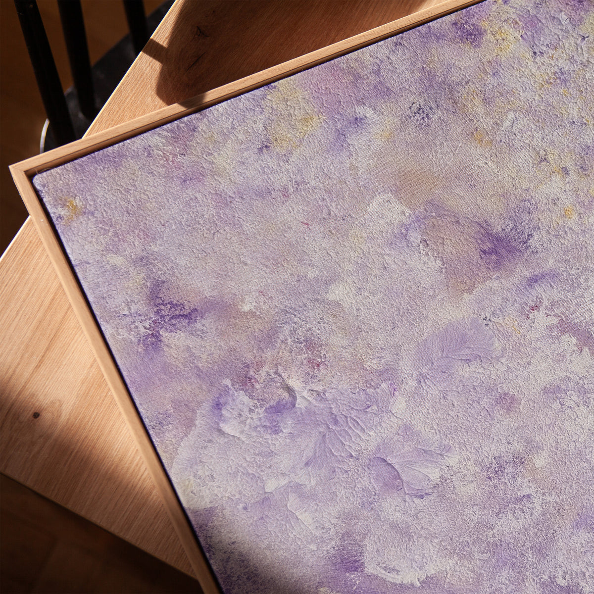 A square canvas painting with a textured, impasto-style surface in shades of lavender purple, white, and soft yellow, displayed on a light wood easel.