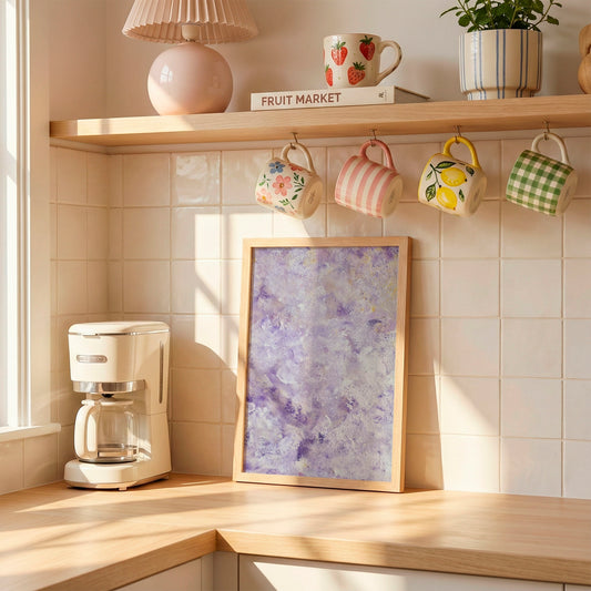 A framed abstract art print with purple and blue brushstrokes leans against a tiled kitchen wall on a wooden counter, next to a white coffee maker. Above, a wooden shelf holds patterned ceramic mugs, a lamp, and a small plant.