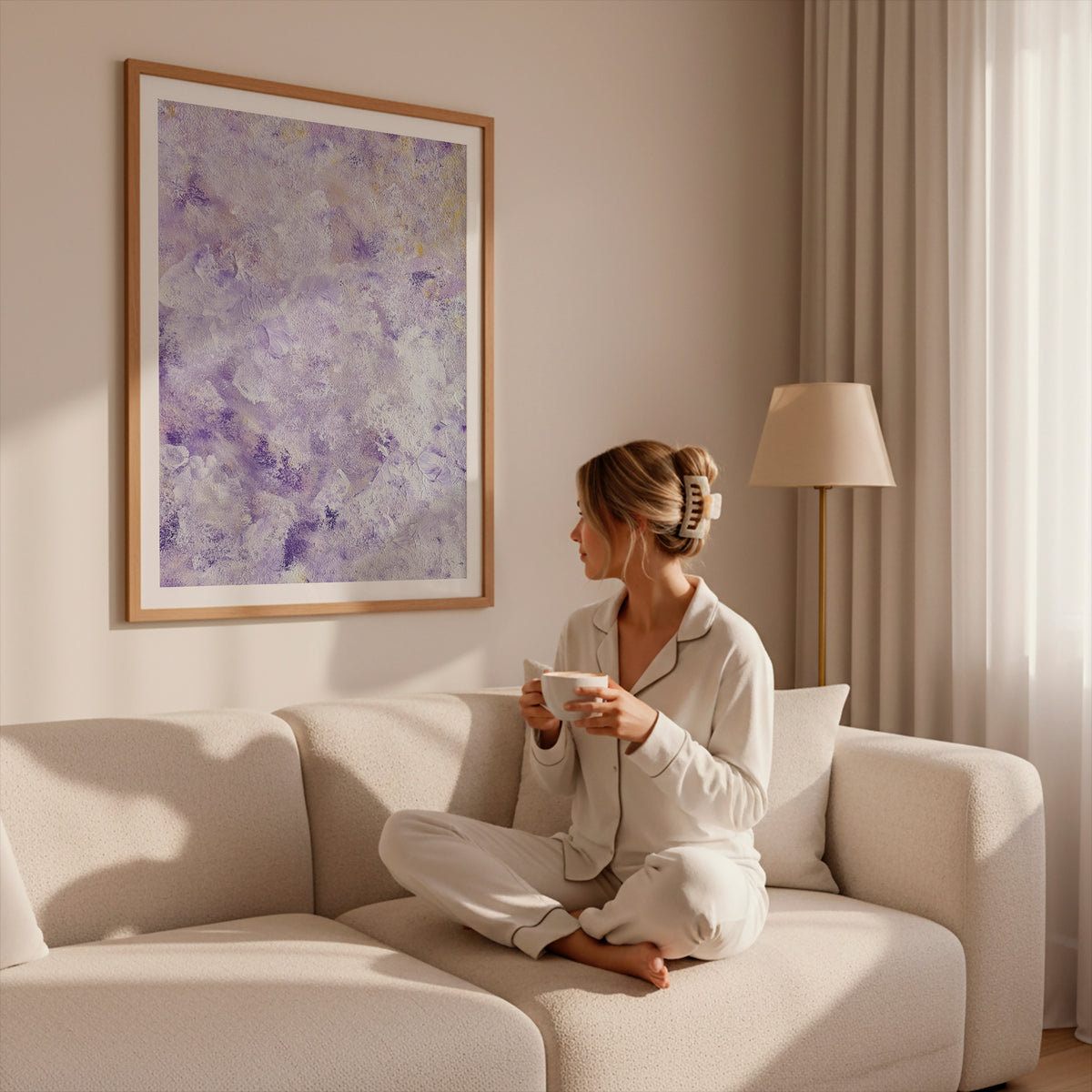 A woman in white pajamas sits cross-legged on a cream sofa in a softly lit living room, holding a white mug. A large framed abstract purple artwork hangs on the wall above her, with a floor lamp and curtains in the background.