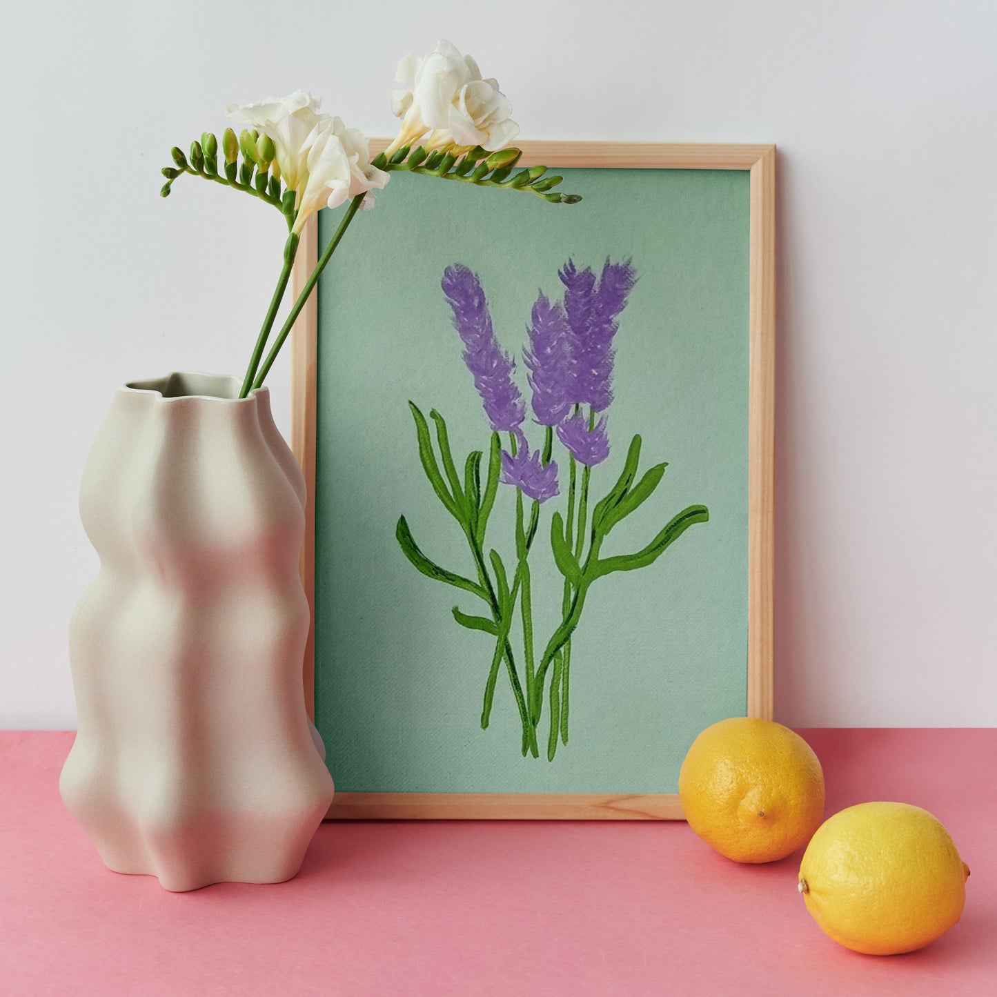 A framed botanical art print of purple lavender on a sage green background, styled on a pink surface next to a beige ceramic vase with white flowers and two yellow lemons against a light gray wall.