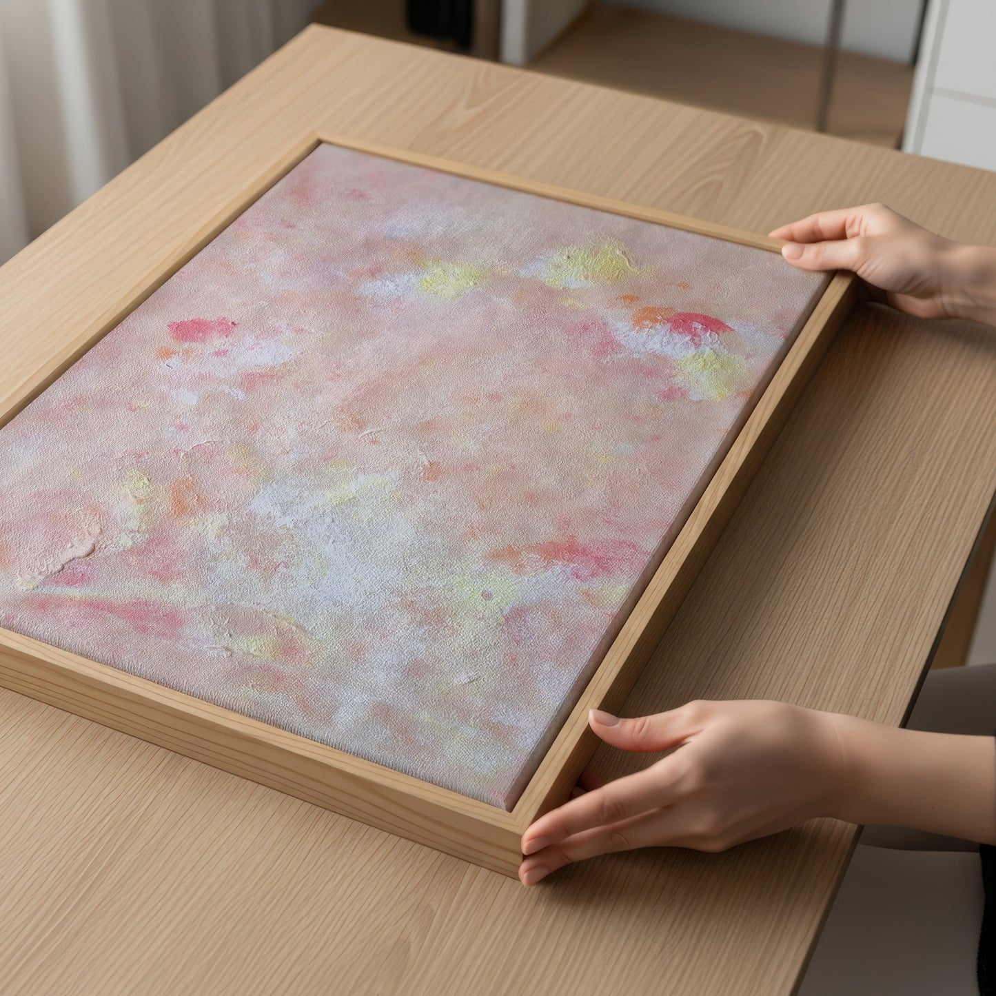 A framed abstract art print with a textured, mottled background of pink, beige, and soft yellow tones, housed in a light natural wood frame. The frame is being held by two hands on a light wood table.