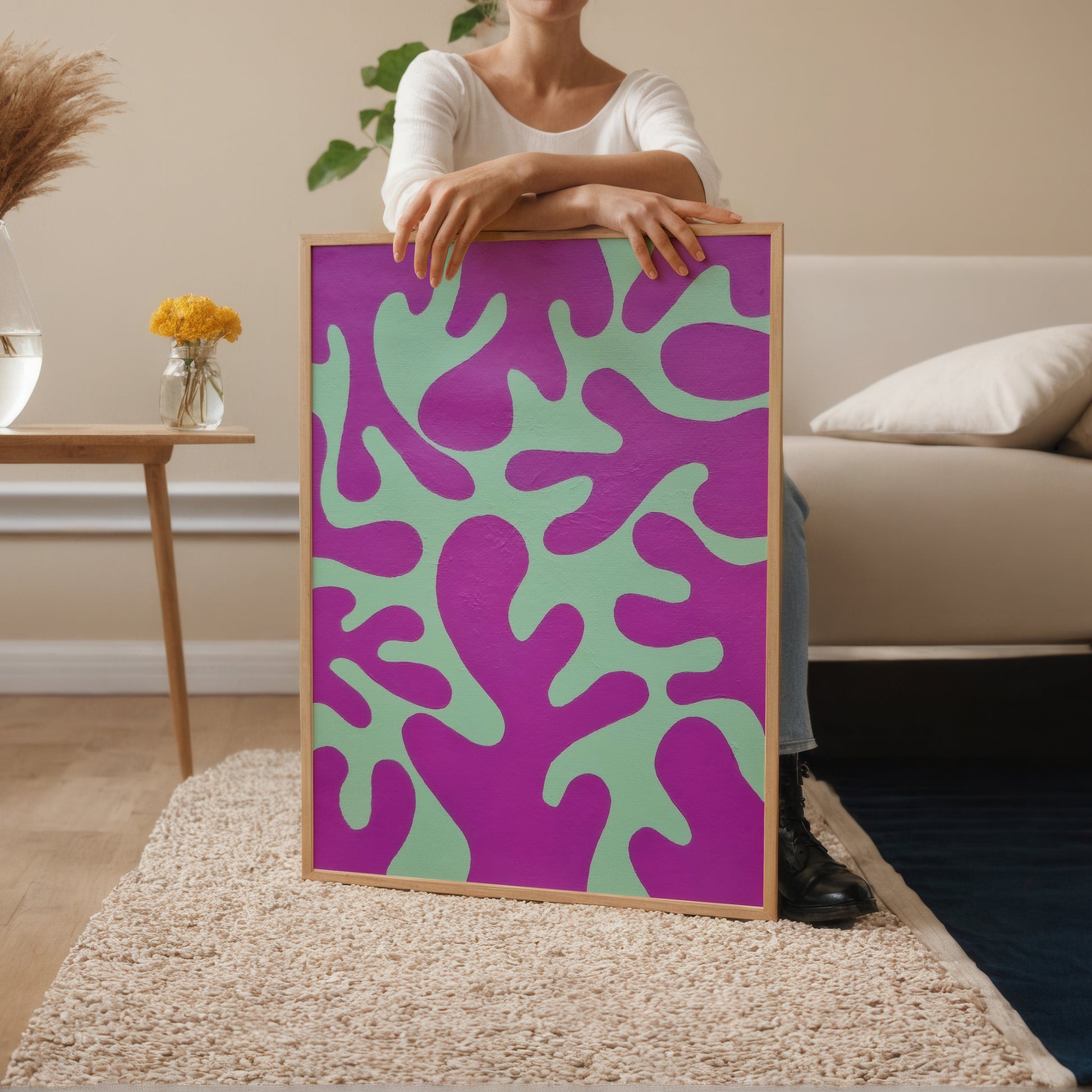 A person in a white top holds a large framed art print featuring a bold abstract pattern in purple and mint green, set in a modern living room with a beige sofa and side table.