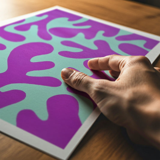 A close-up of a hand touching a square art print with a bold abstract pattern in purple and mint green, resting on a wooden surface.