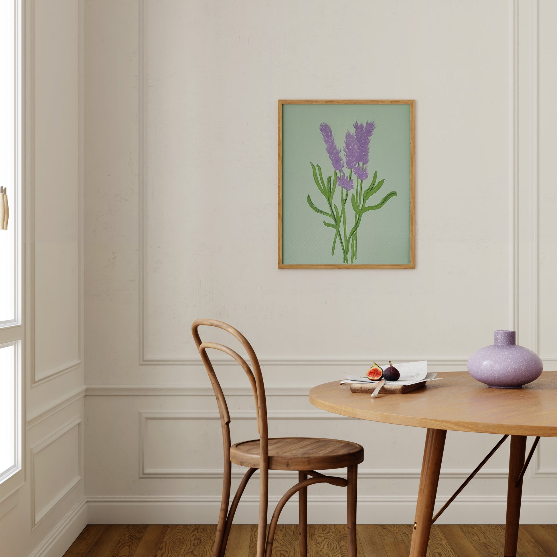 A framed botanical art print of purple lavender flowers on a sage green background, hanging on a cream-colored wall above a wooden dining table and chair.