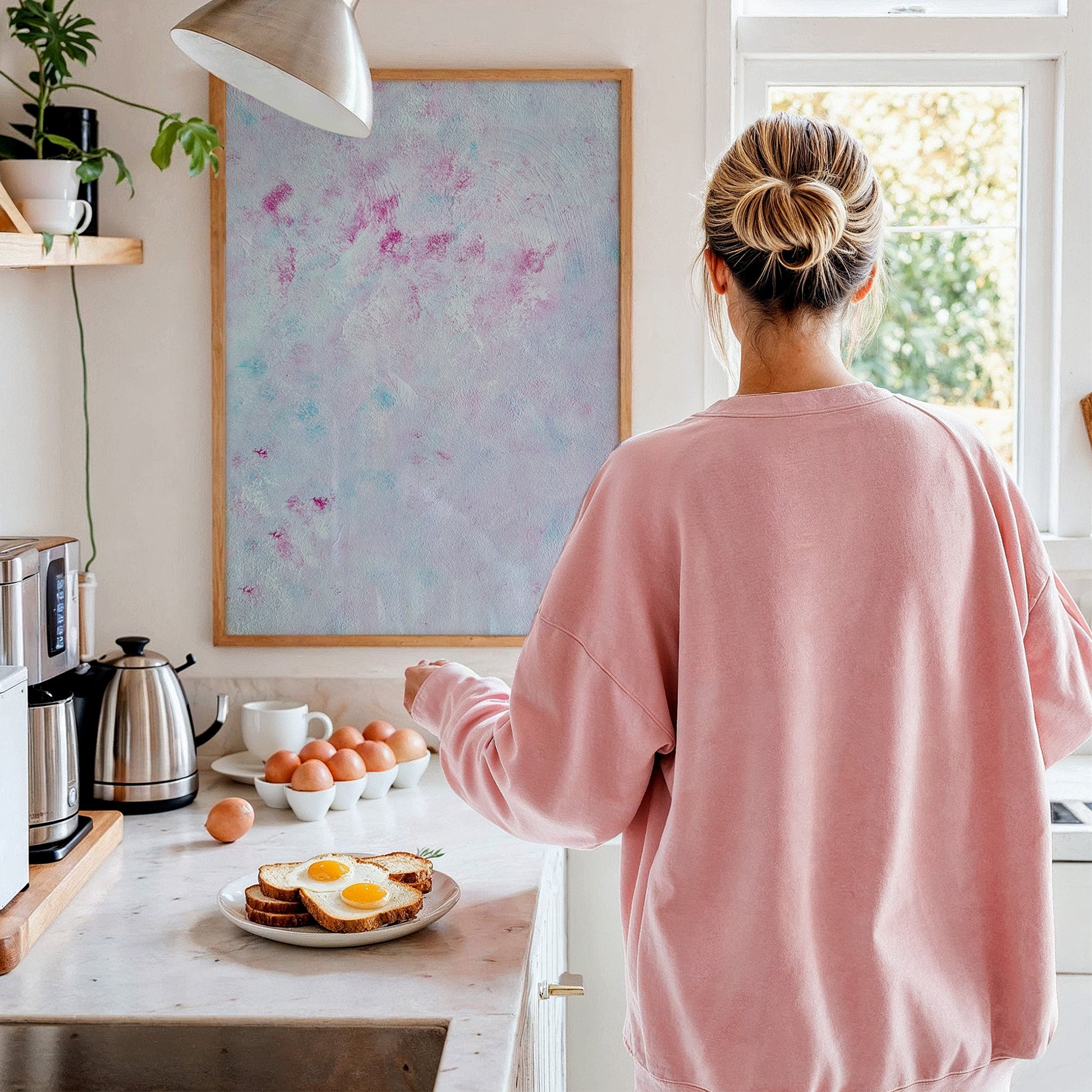 A woman with her hair in a bun stands with her back to the camera in a bright kitchen, wearing an oversized light pink sweatshirt. On the marble countertop are eggs, a plate with toast and fried eggs, a kettle, and a large framed abstract painting on the wall.