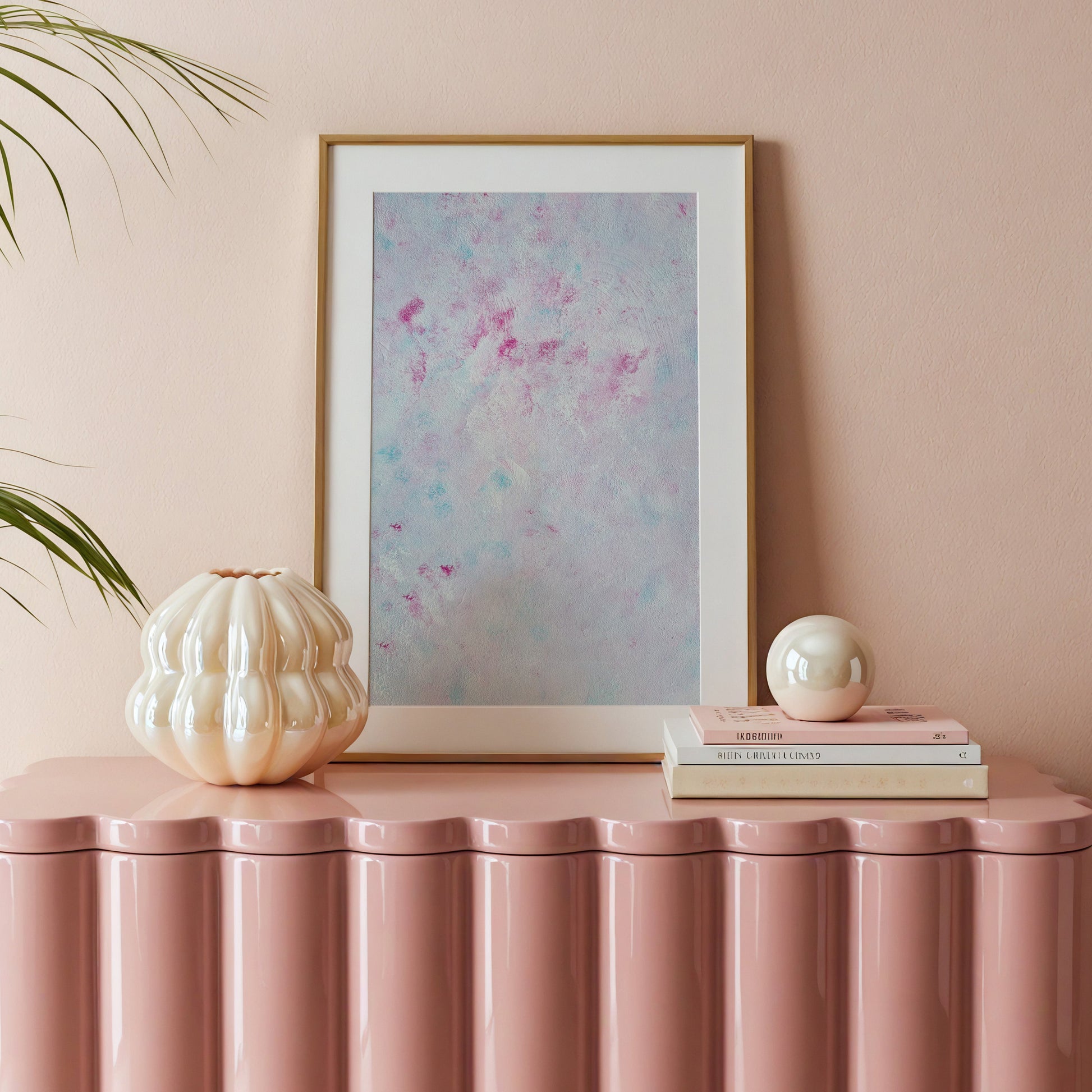 A framed abstract art print with a gold frame and white mat, featuring a pastel blue and pink watercolor design, styled on a pink ribbed console table with decorative objects against a matching pink wall.