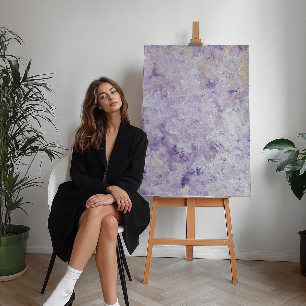 A woman in a black blazer sits on a white chair next to a wooden easel holding a textured purple and lavender abstract painting, with potted plants on a light wood herringbone floor against a white wall.