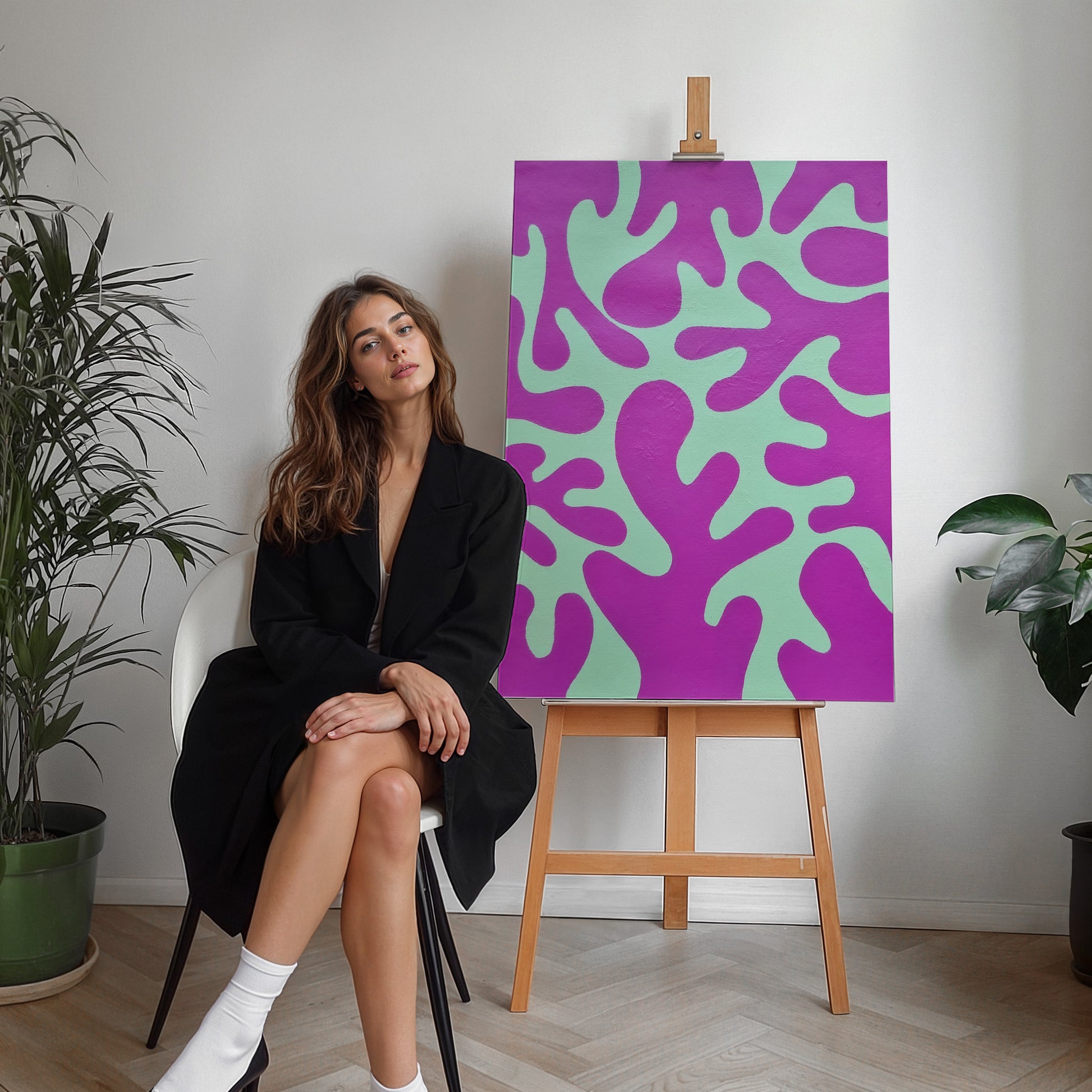 A woman in a black blazer sits on a white chair next to a canvas print on a wooden easel. The canvas features an abstract organic pattern in bright purple and mint green.