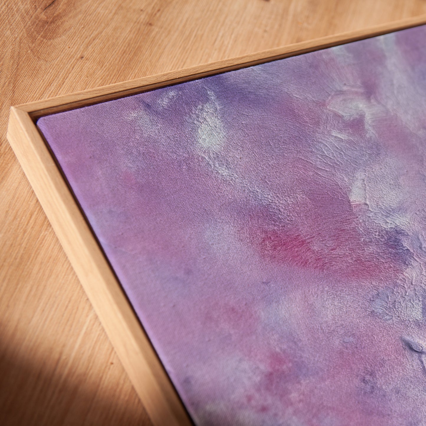A close-up of a framed abstract art print featuring textured purple, pink, and lavender tones, set in a light natural wood frame on a wooden tabletop.