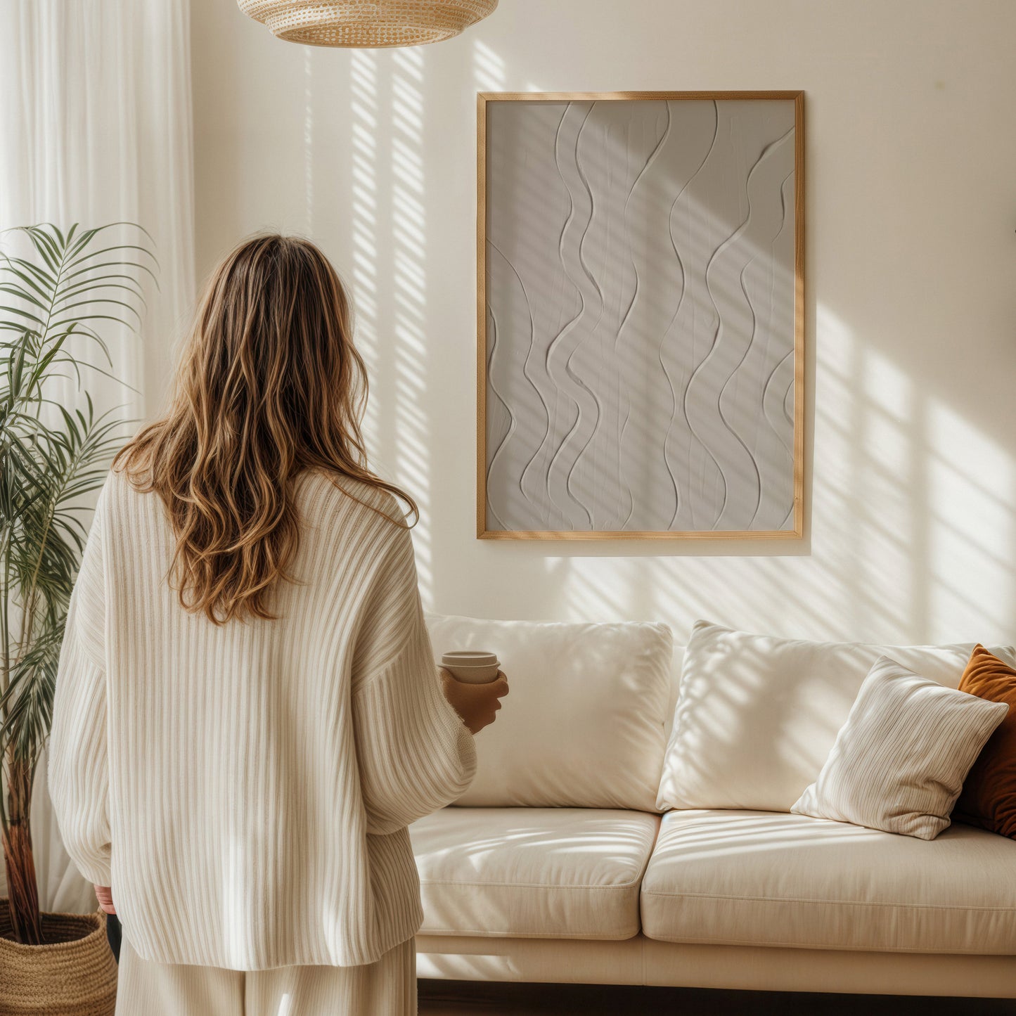 A woman stands with her back to the camera in a bright, sunlit living room, wearing a cream ribbed cardigan and matching pants. She is positioned in front of a cream sofa with decorative pillows and a framed abstract wall art piece.