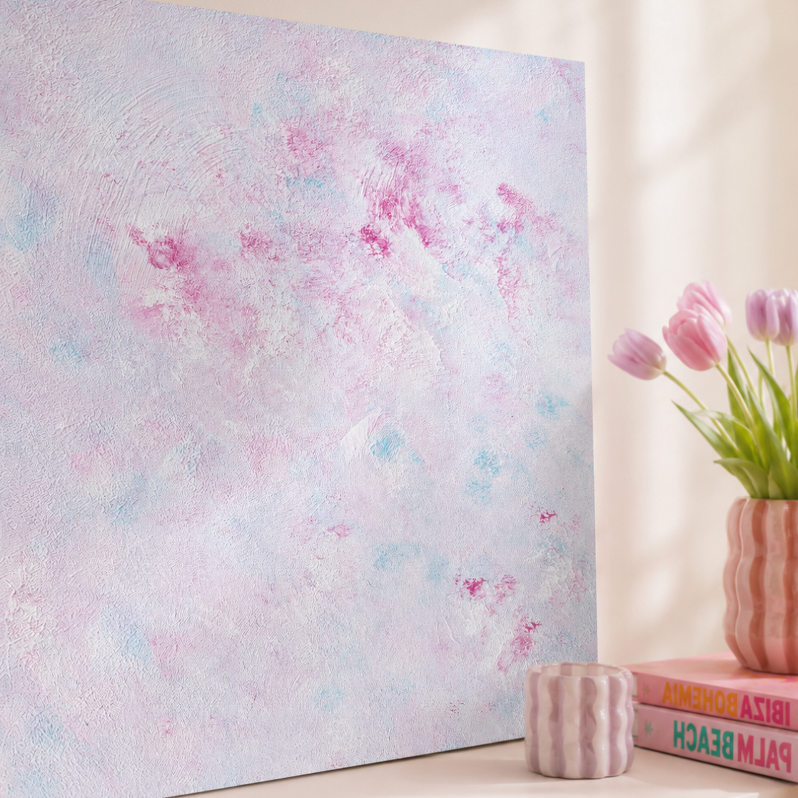 A square canvas wall art print with a textured abstract design in pastel pink, lavender, and light blue, displayed leaning against a beige wall on a white shelf next to decorative props.