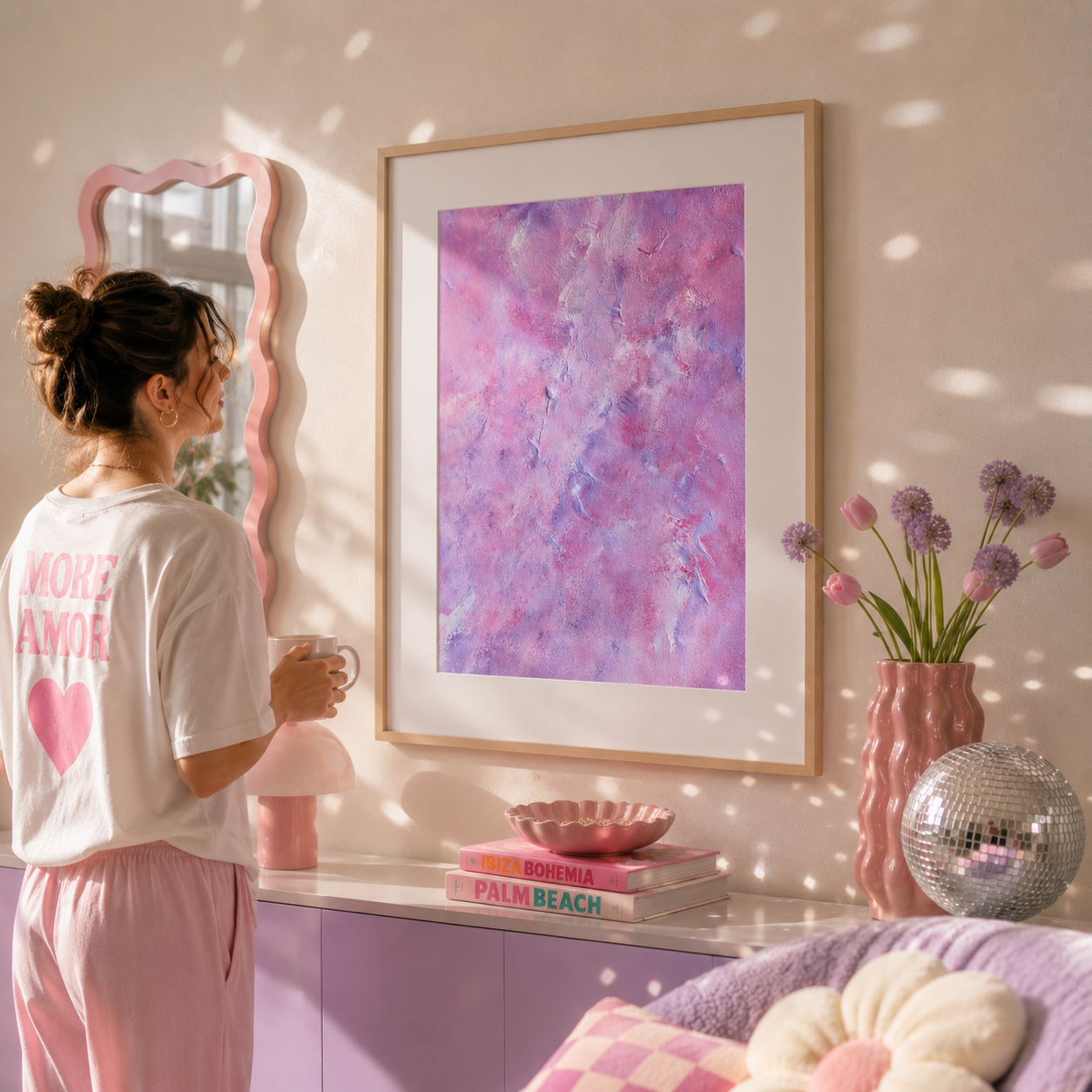 A woman in a white t-shirt and pink pants stands in a sunlit room looking at a large framed abstract art print with pink and purple tones. The room has a pink mirror, a pink lamp, a vase of flowers, and a purple cabinet.