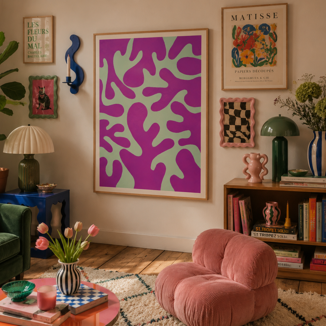 A cozy, retro-styled living room featuring a large framed abstract art print with magenta and mint green shapes on the wall. The room is decorated with a green sofa, a pink velvet armchair, a bookshelf, and various plants and decorative items.