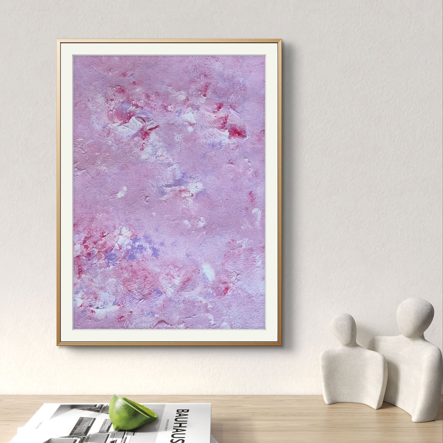 Original abstract acrylic art piece in pink, lilac and red, hanging vertically against a beige wall over a desk with magazines, ceramic sculptures and a green apple