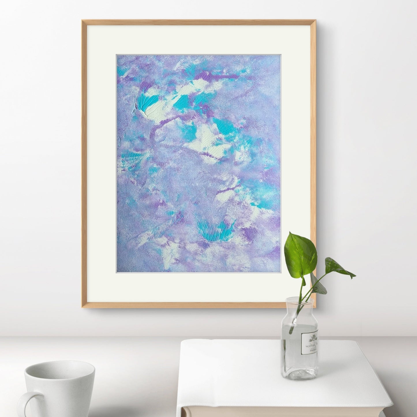 Original abstract acrylic colorful art piece in pastel purple, blue and white hanging vertically as decor against a white wall over a white modern desk with a minimalistic ocffee table book, a green plant and a coffee cup