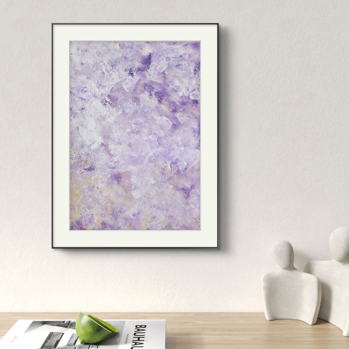 Original abstract colorful acrylic art piece in deep purple and pastel yellow hanging vertically on a white wall over a light oak minimalistic desk over interior magazines and light stone figures