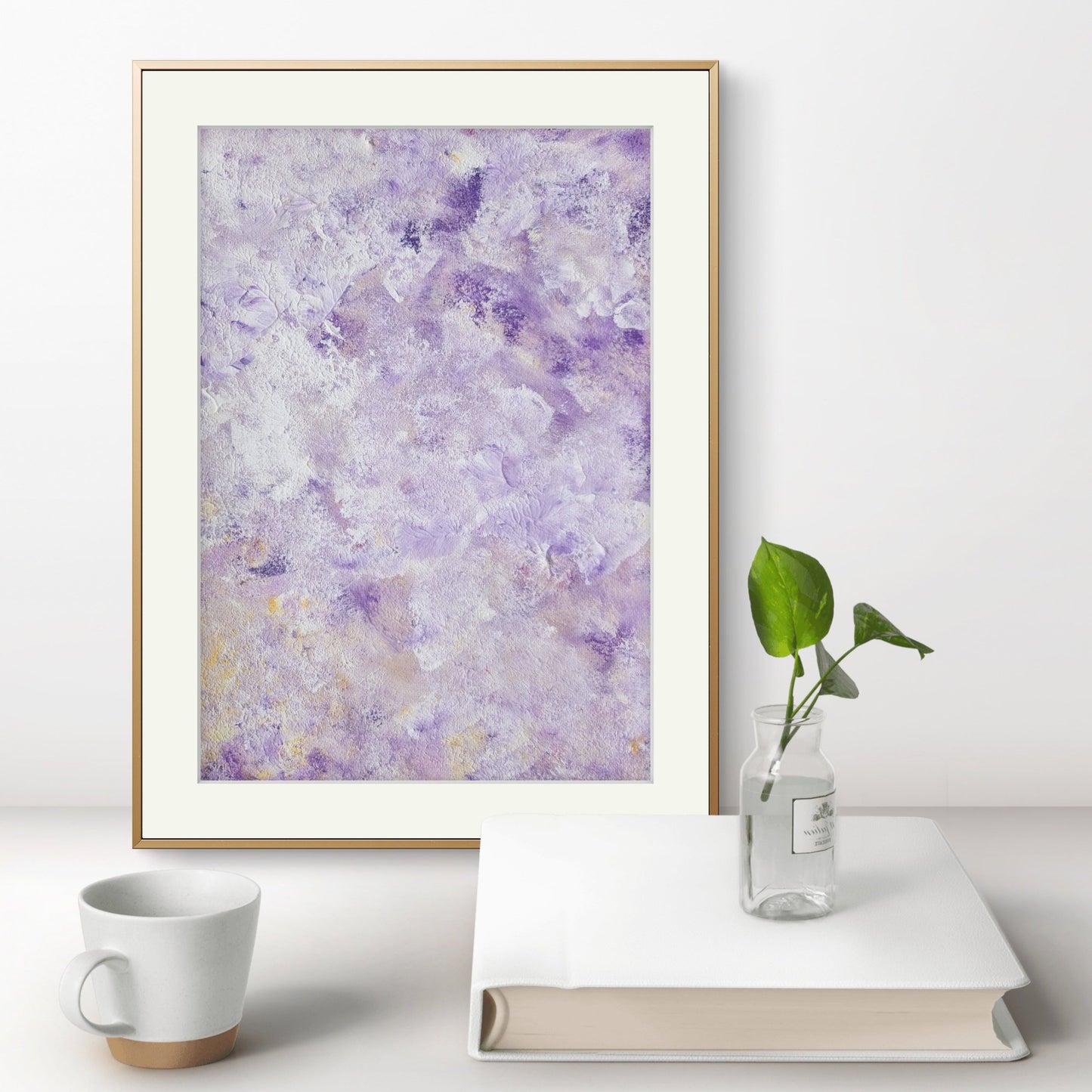 Original abstract colorful acrylic art piece in deep purple and pastel yellow leaning vertically on a white wall on a modern minimalistic white desk behind a white coffee table book, a green plant and a white coffee mug