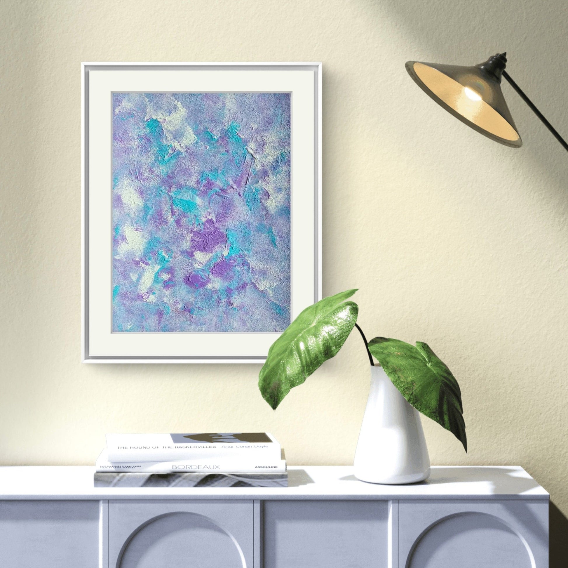 Original one of a kind colorful abstract acrylic art piece in pastel purple, blue and white, hanging vertically against a beige wall in a modern home over a blue dresser with green leaves, minimalistic coffee table book with a black designer lamp