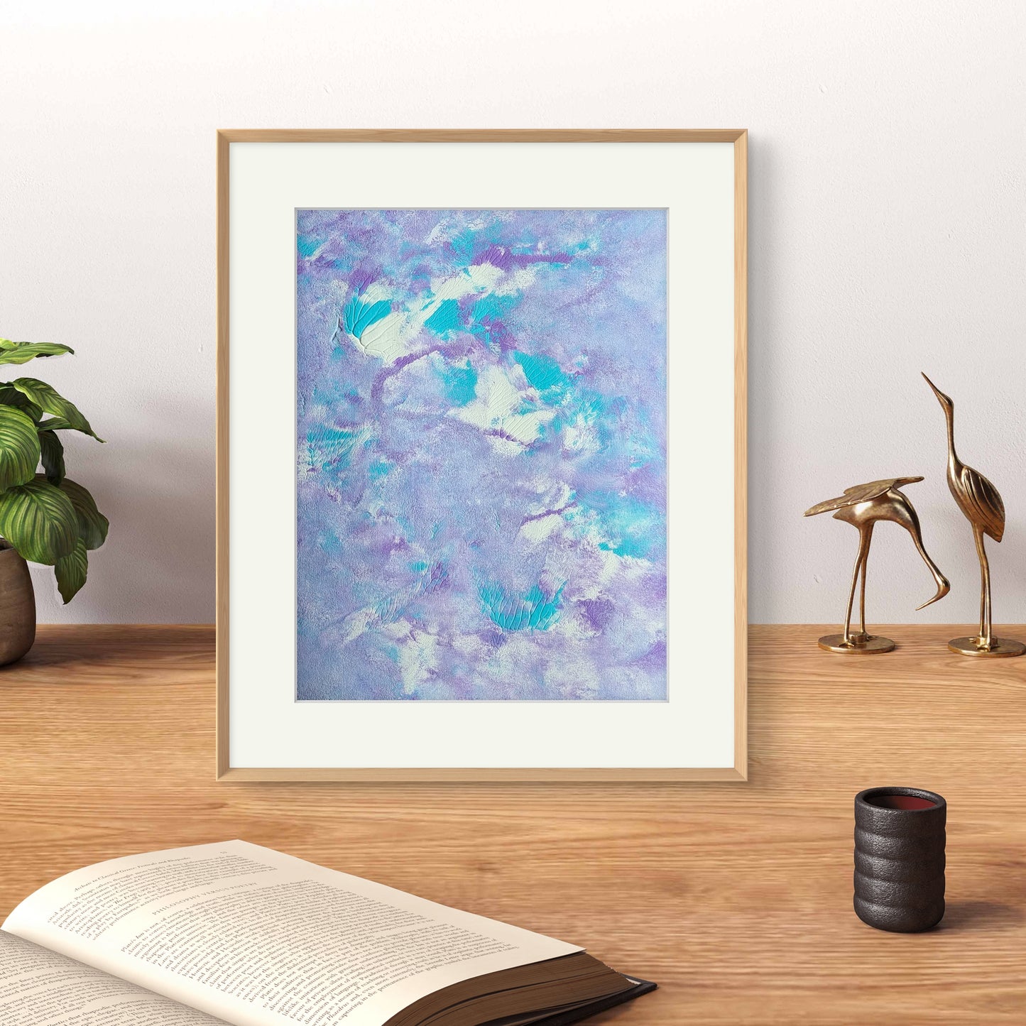 Original one of a kind colorful abstract acrylic art piece in pastel purple, blue and white, leaning vertically against a white background as decor on top of an oak desk together with an open book, brass decor figures and a green house plant