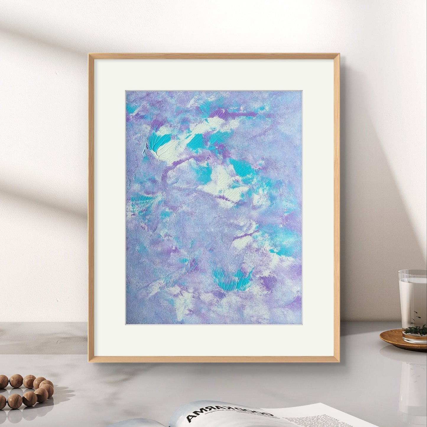 Original one of a kind colorful abstract acrylic art piece in pastel purple, blue and white, leaning vertically against a white whall as decor on top of a white marmor desk with a decor magazine and sented candle