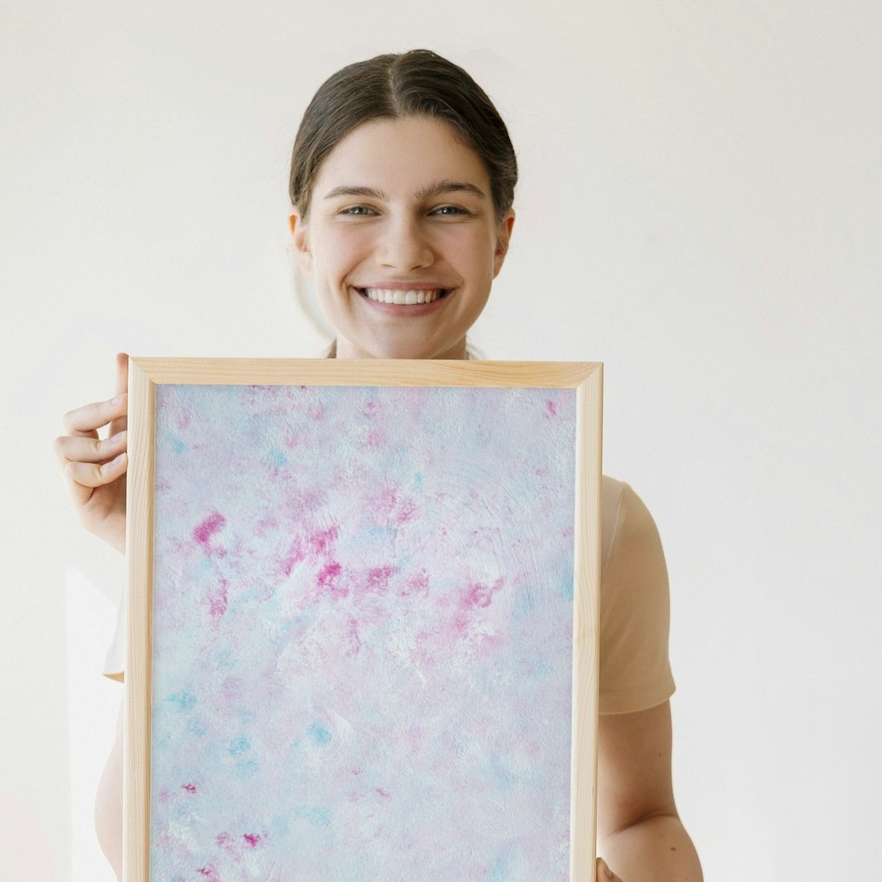Pastel blue, light pink, red and white abstract art poster in a light oak frame, held in the hands of a smiling woman with brown hair in a pony tail wearing white and beige clothing. The wall behind the poster and woman is eggshell white. 