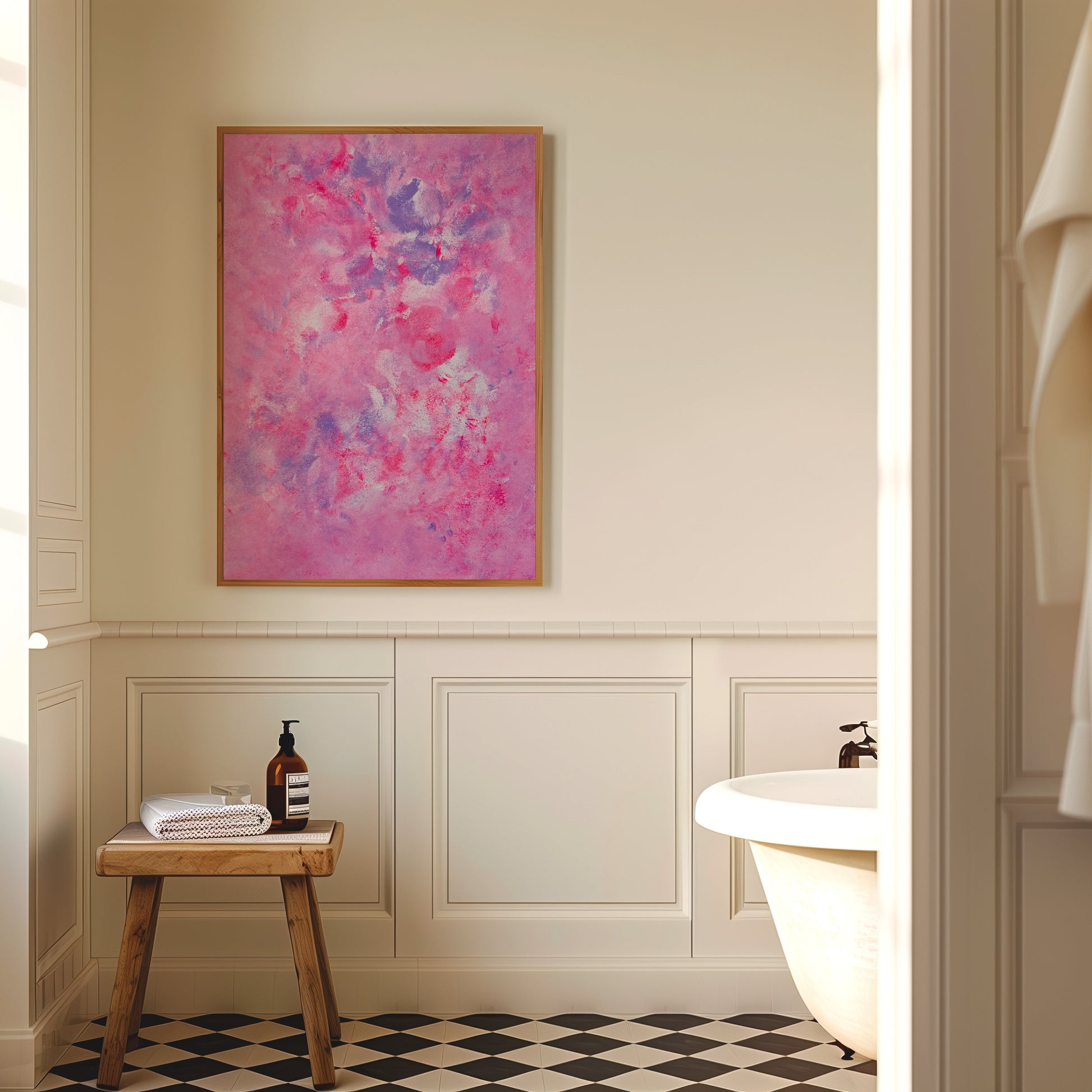A framed abstract painting with vibrant pink, purple, and blue brushstrokes hangs on a cream-colored bathroom wall above wainscoting, next to a small wooden stool and a white bathtub.